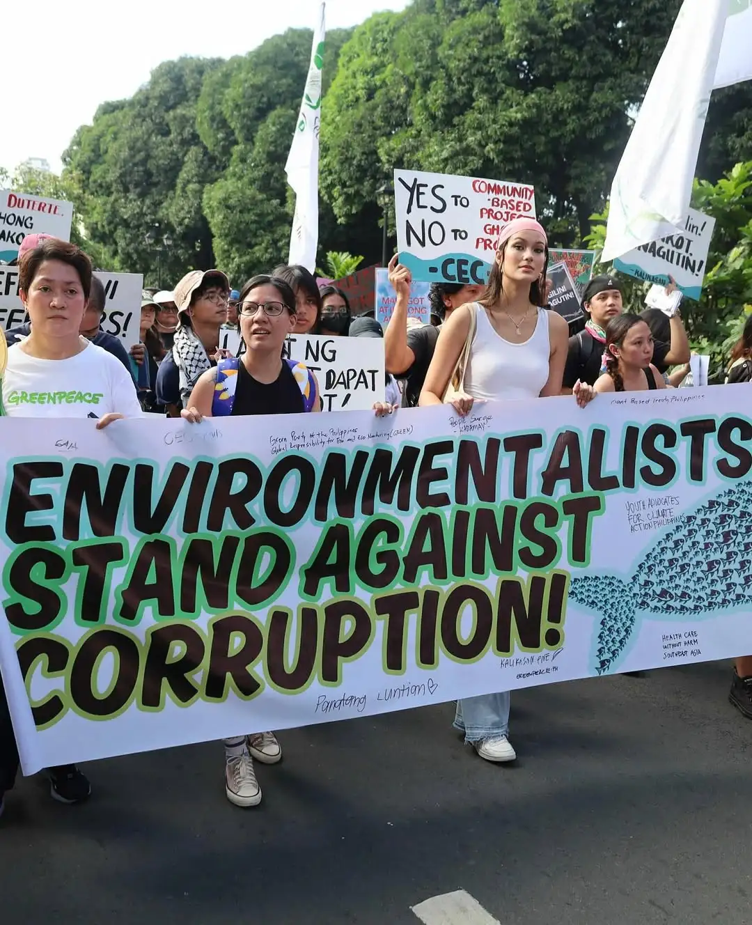Protesters hold a large banner reading 'ENVIRONMENTALISTS STAND AGAINST CORRUPTION!' during a street demonstration, with people behind them carrying signs.
