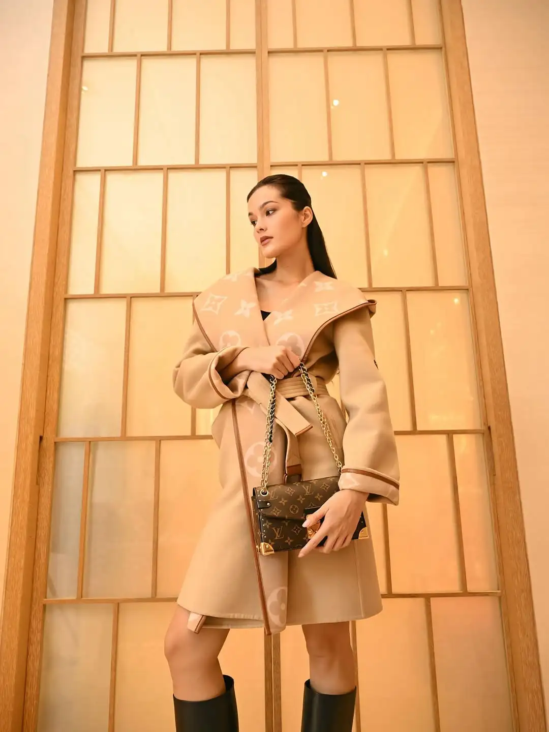Woman in a beige oversized coat posing with a Louis Vuitton handbag against a wooden paneled wall.