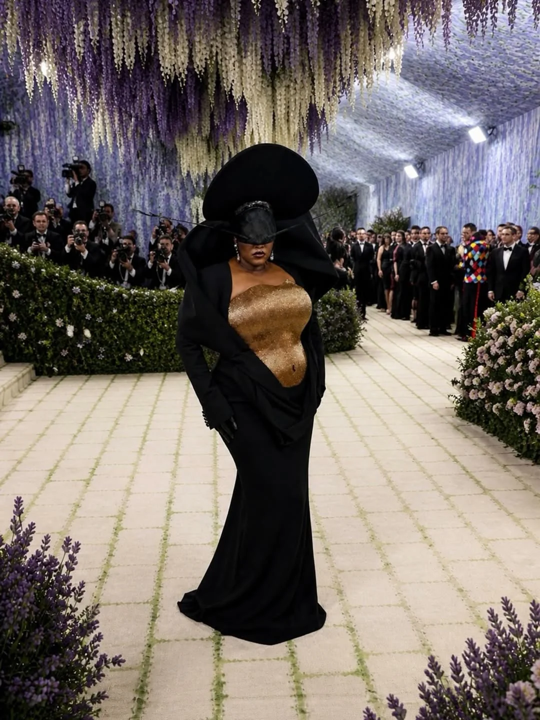 Model in a gold sequined bodice and dramatic black cape with a huge hat walks a flower-decked runway while photographers look on.