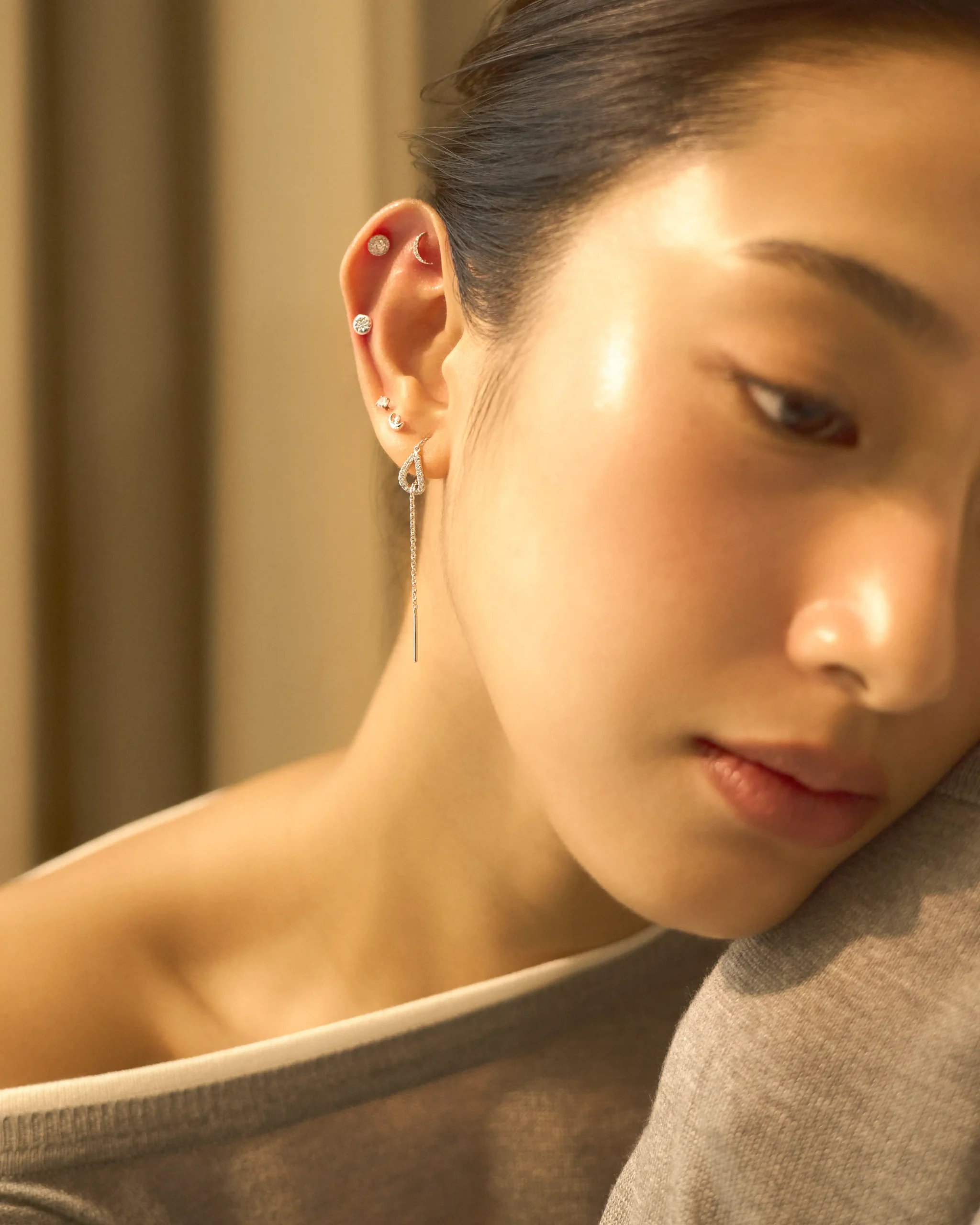 Close-up of a ear with multiple silver diamond studs, a crescent moon cuff, and a long dangling earring against a warm background. All Day, All Night: Suki Jewelry Tells Time Differently