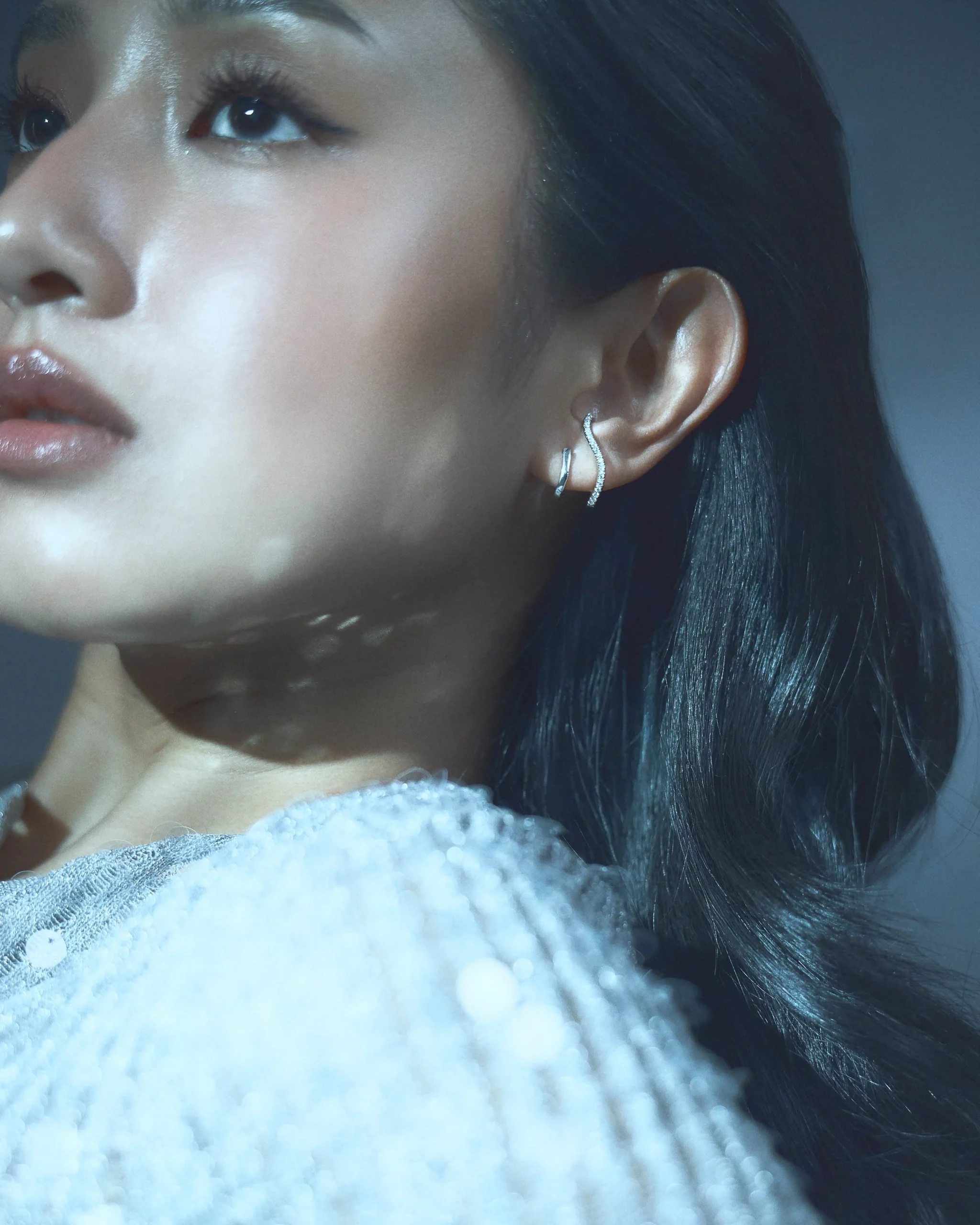 Close-up of a woman’s profile: visible eye and glossy lips, curved silver earrings, with a white fuzzy scarf in the foreground.