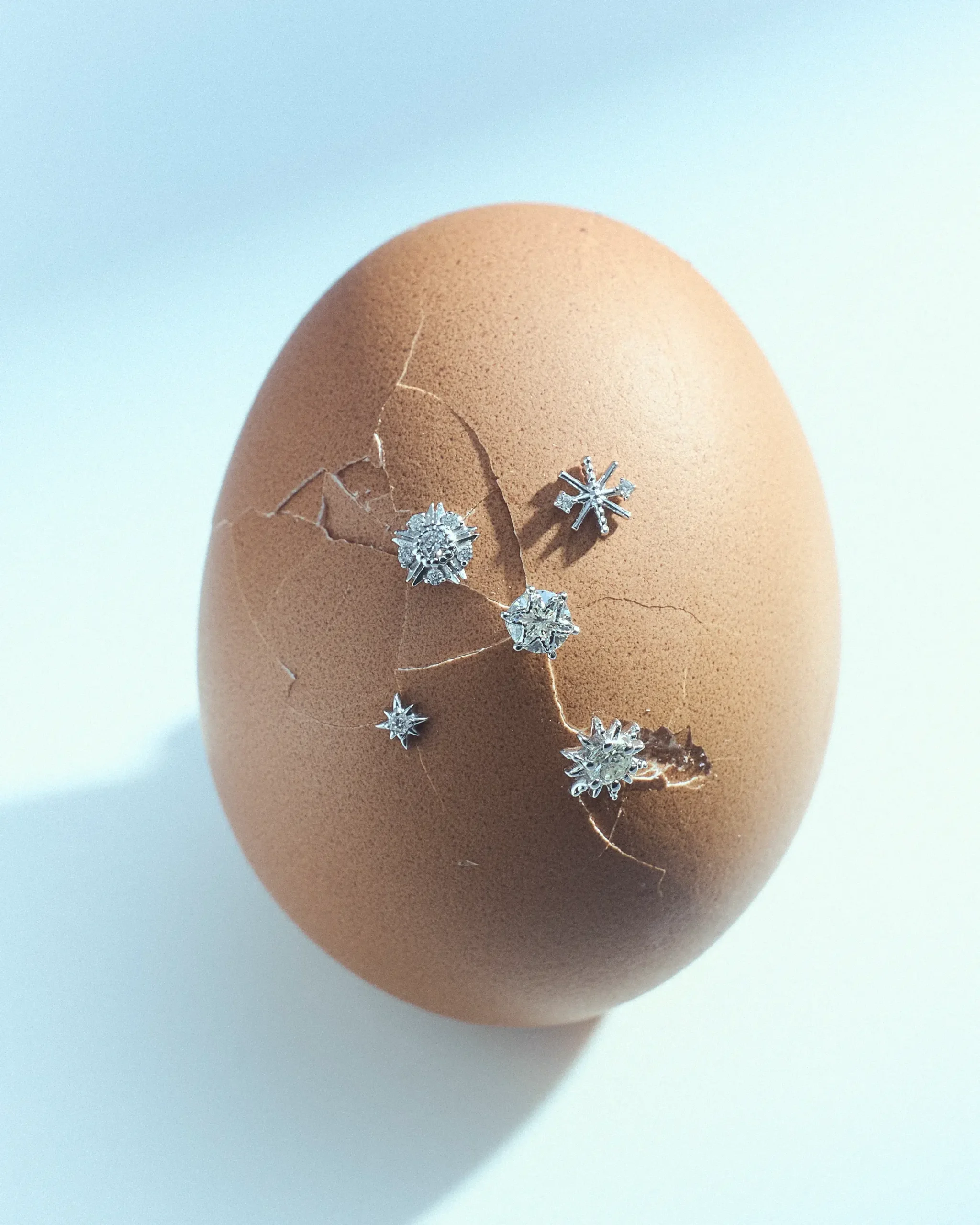 Close-up of a brown eggshell with fine cracks, wrapped by small silver snowflake jewelry charms resting on its surface.
