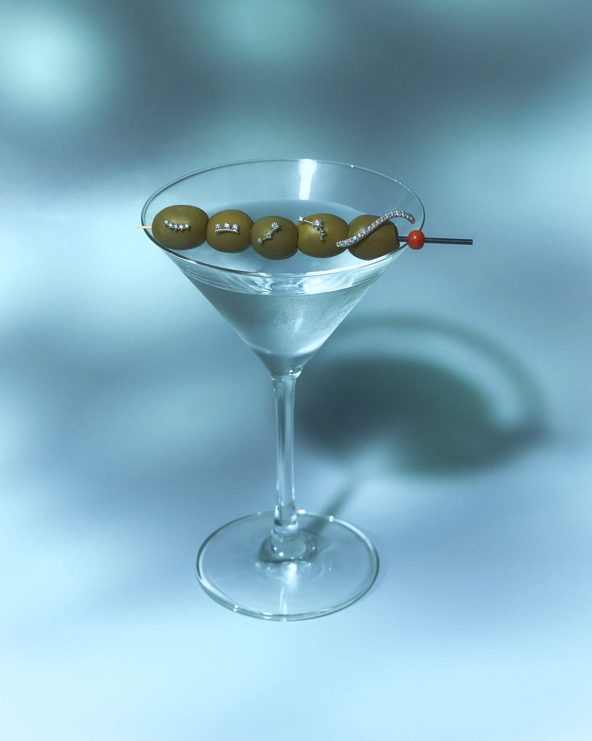 Martini glass with a skewer of five olives, each olive wearing tiny diamonds; red bead at the end on a blue background.