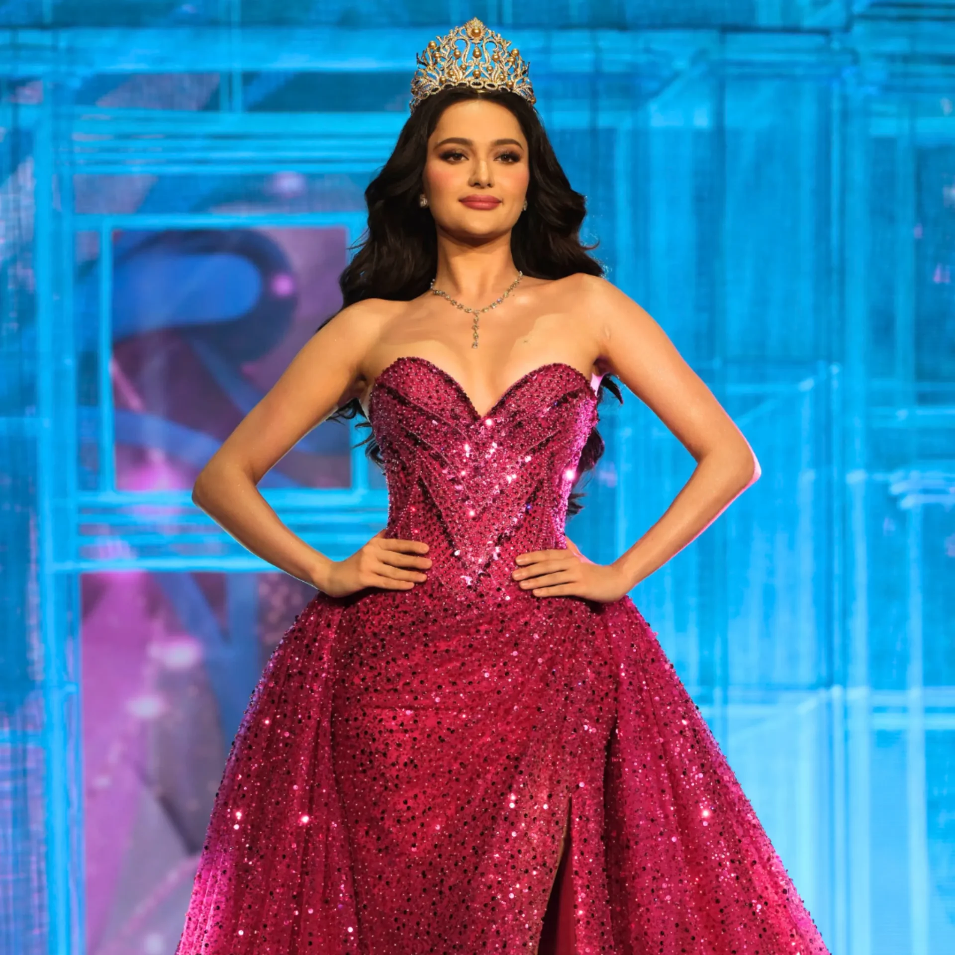 Woman wearing a jeweled crown and a pink sequined gown posing on a blue-lit stage backdrop.