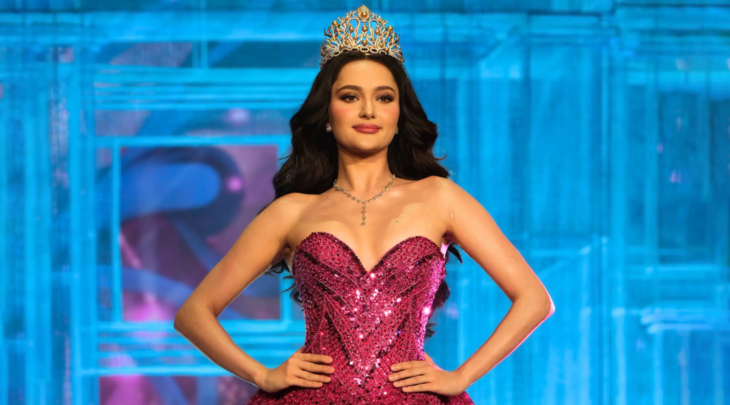 Woman in a jeweled crown and pink sequined gown on a blue stage, posing with hands on hips.