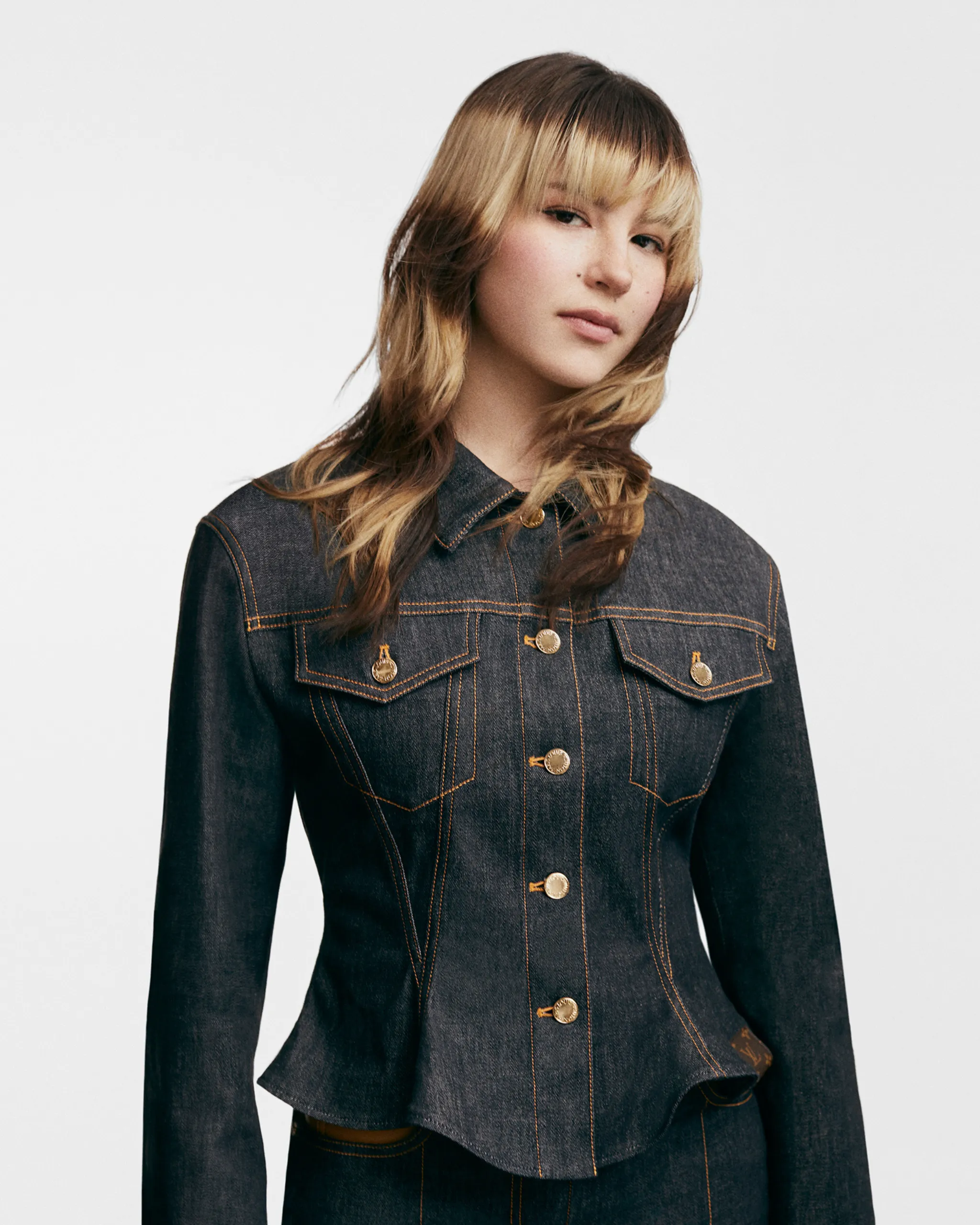 Young woman in a dark denim jacket with orange stitching, facing slightly to the right against a light gray background.