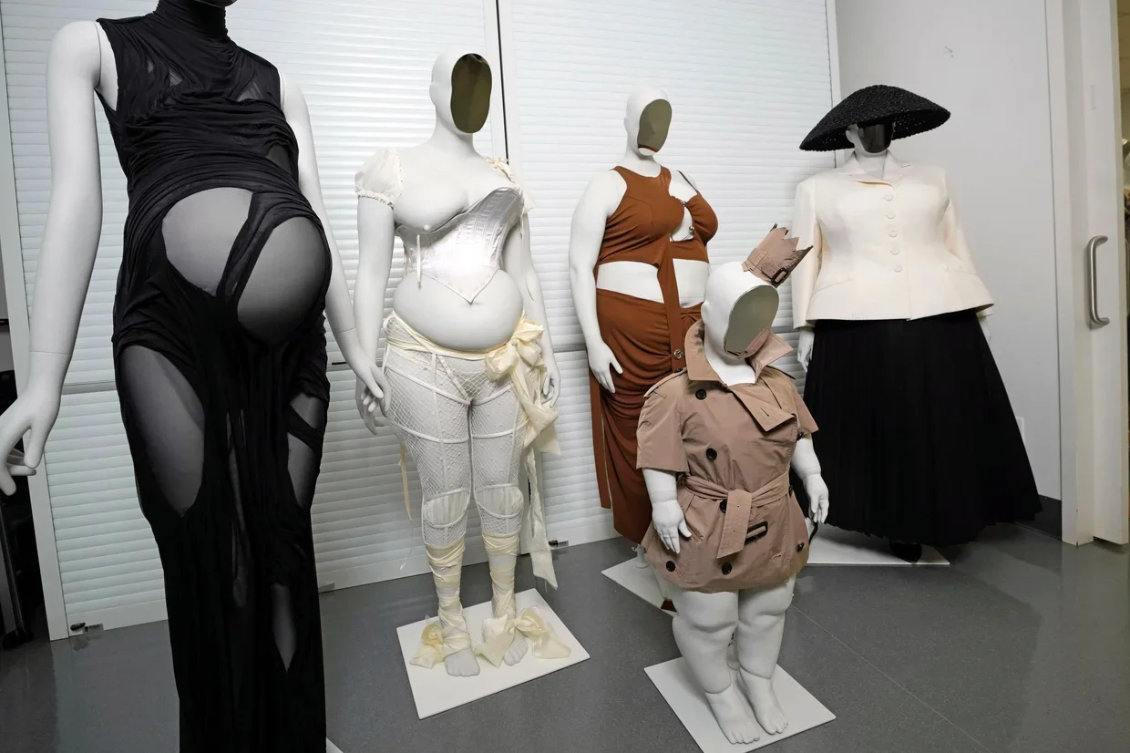 Mannequins in a fashion display wearing a range of outfits: a black sheer gown with cutouts, a white corseted bodice and pants, a rust-brown fitted dress, a child-sized beige trench with belt, and a white jacket with a black skirt and a large black hat.