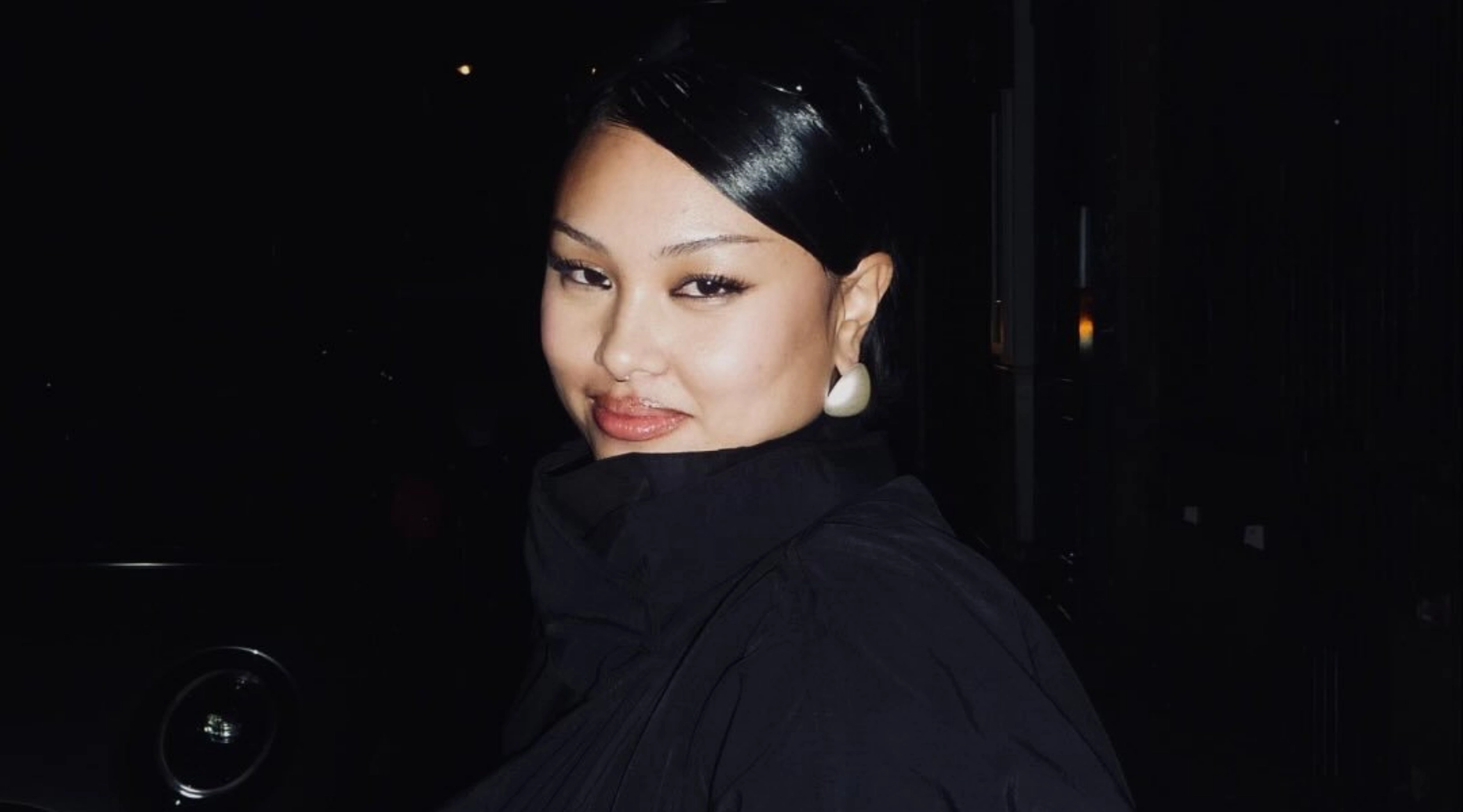 Close-up portrait of a woman with black hair, bold makeup, and a white earring against a dark background.