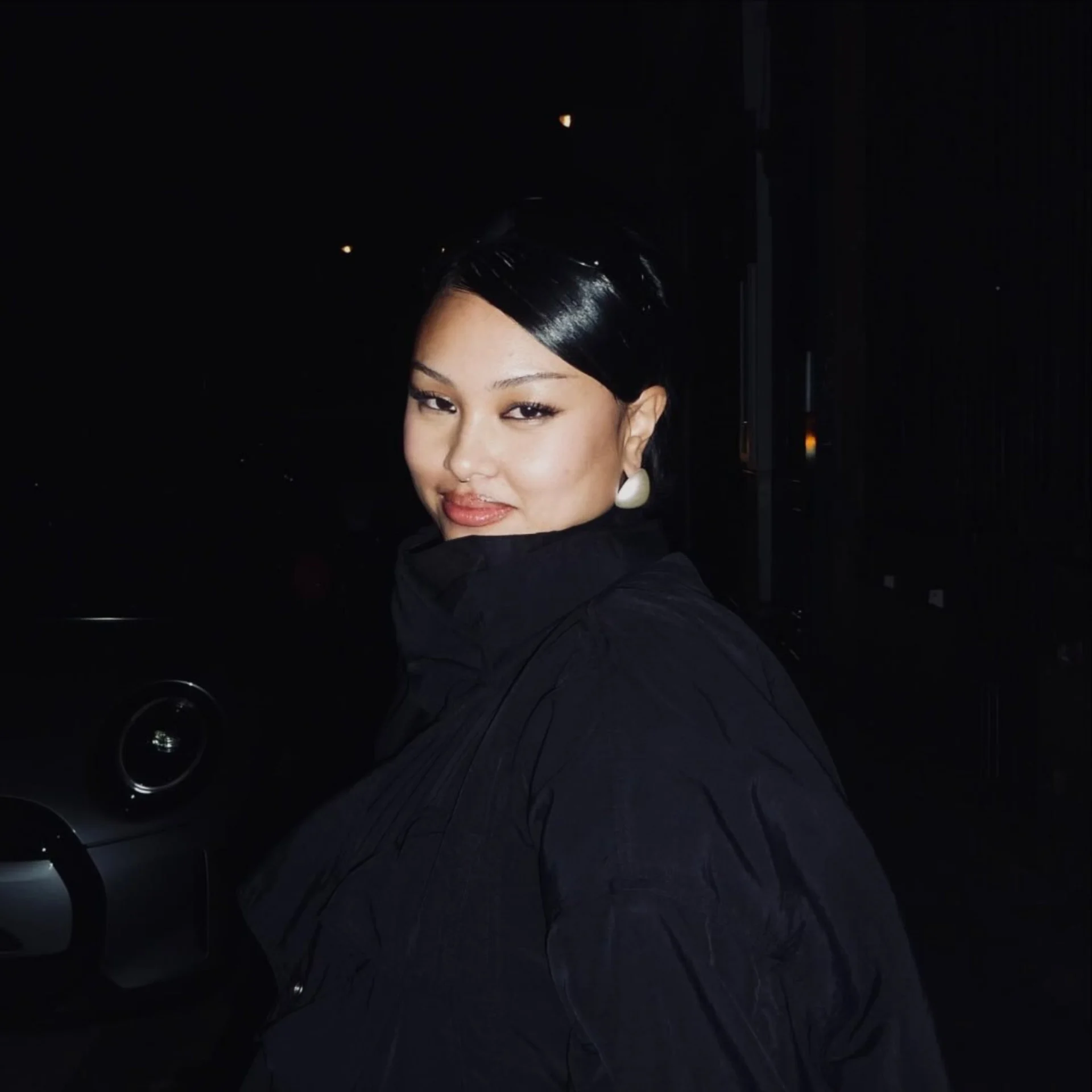 Close-up portrait of a woman in a dark car at night, her face softly lit and looking toward the camera, wearing white earrings and a dark jacket.