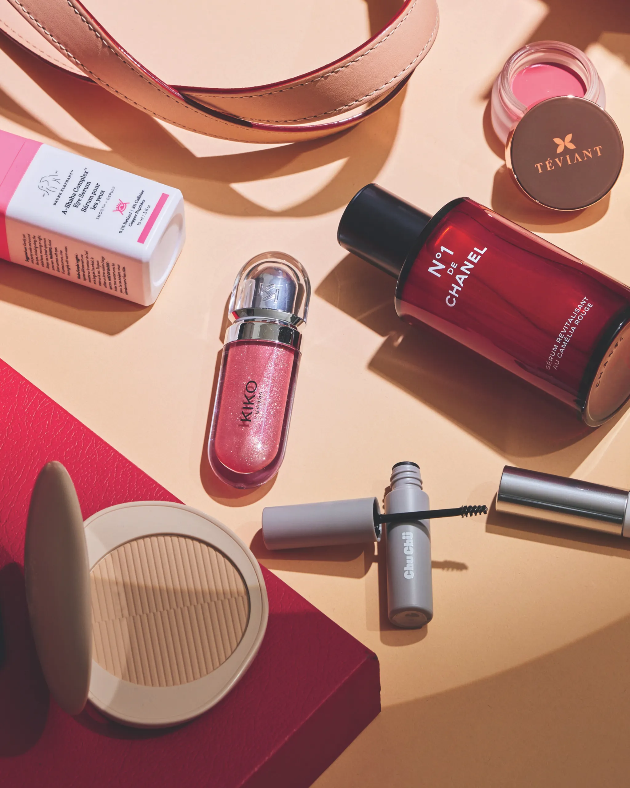 Flat lay of beauty products on a peach surface: Chanel No. 7 perfume bottle, pink lip gloss, Teviant jar, and other pink makeup items.