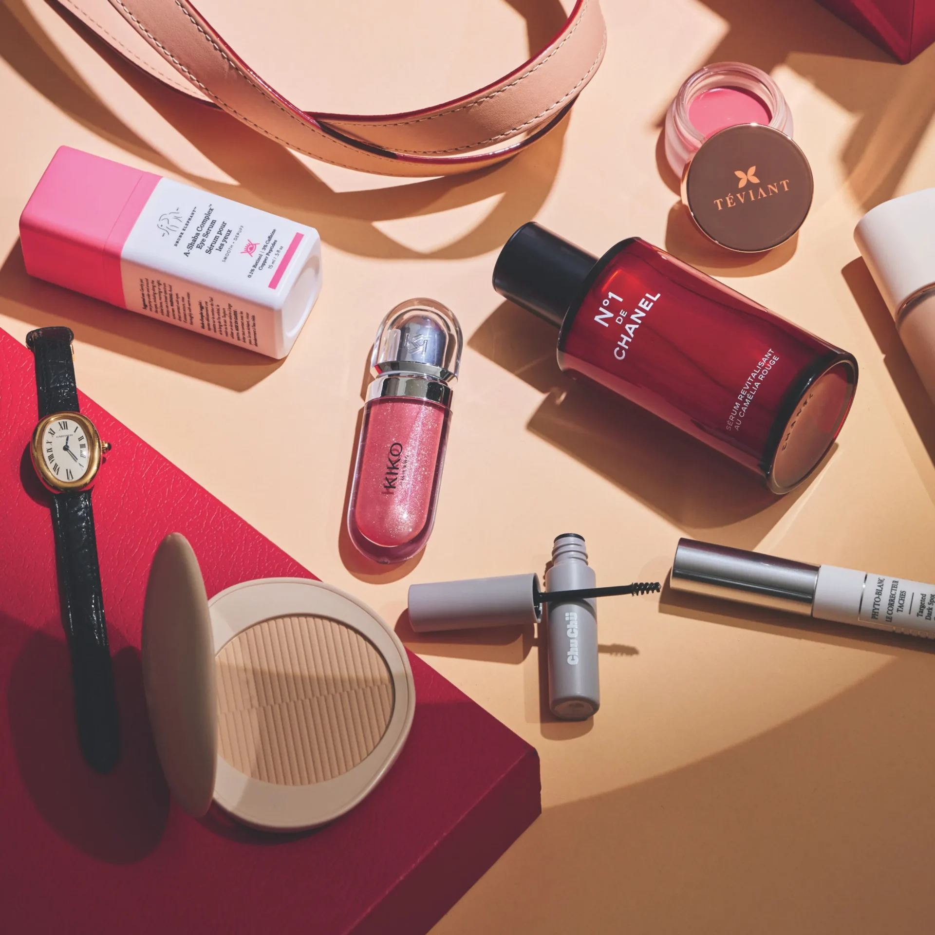 Assorted makeup and accessories spread on a peach surface: pink lipstick, compact powder, a pink belt, a small gold-toned watch, and a bottle labeled Chanel No. 1 along with Téviant makeup case.