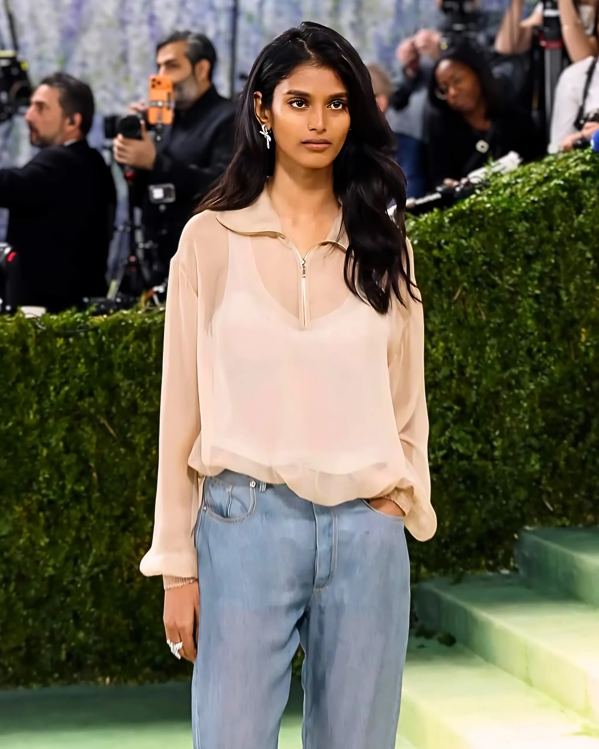 Woman posing at an outdoor event with photographers in the background, wearing a pale beige blouse and light-wash jeans.
