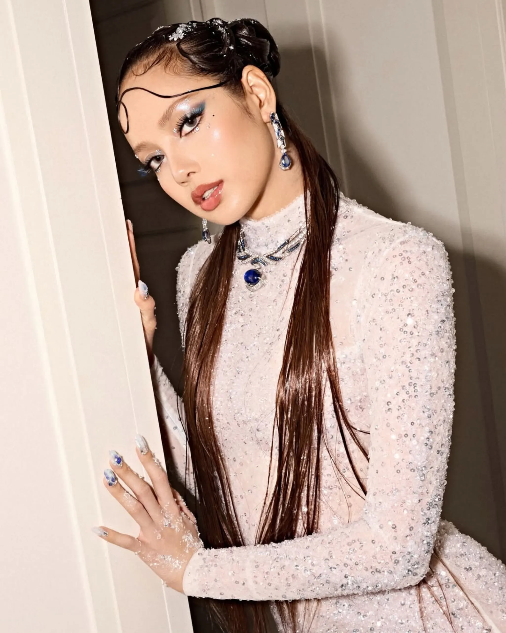 Glamorous woman in a sequined white gown leaning against a doorframe, wearing blue jewelry and bold makeup.