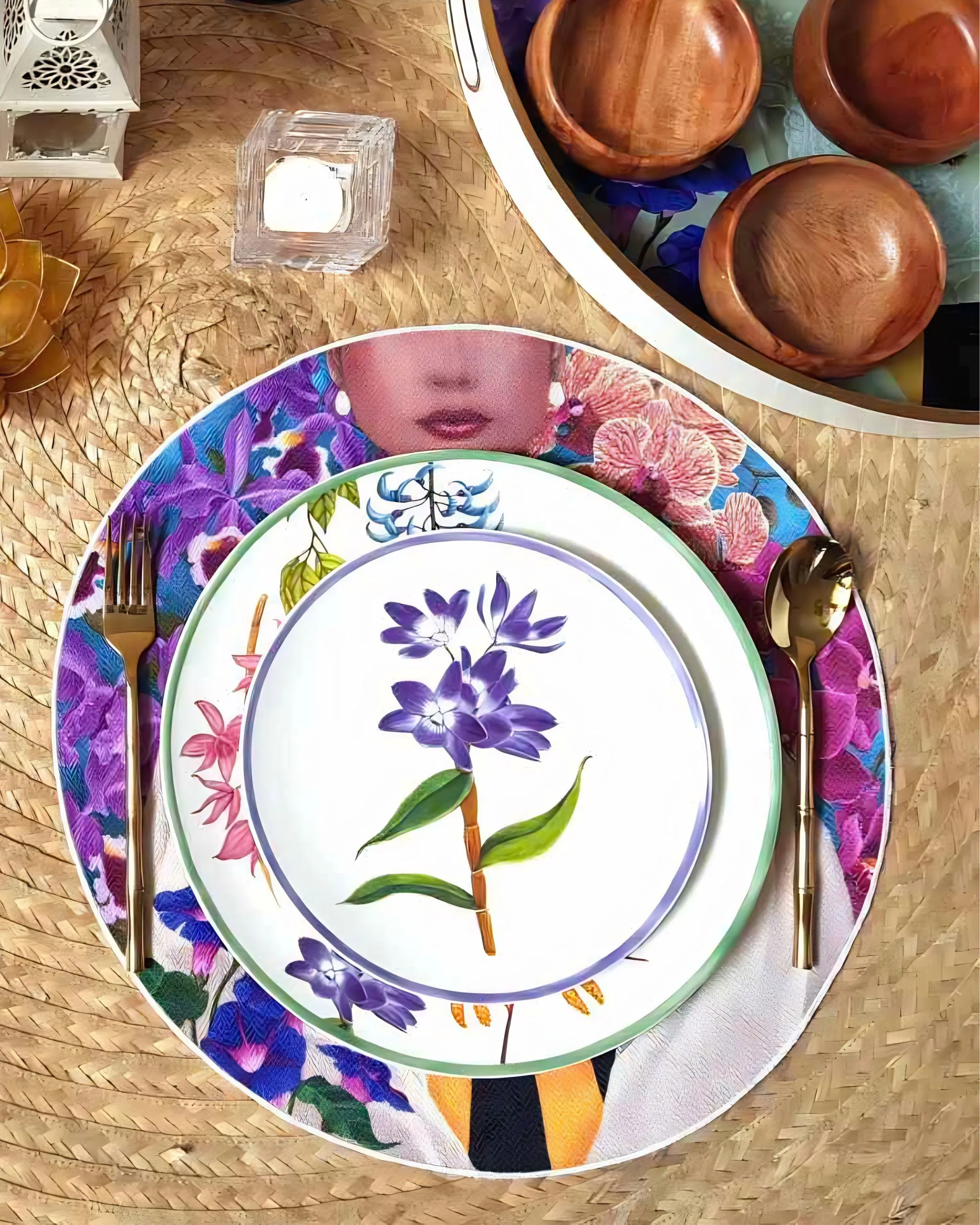 Top-down view of stacked floral plates on a woven mat, with wooden bowls and gold cutlery nearby.