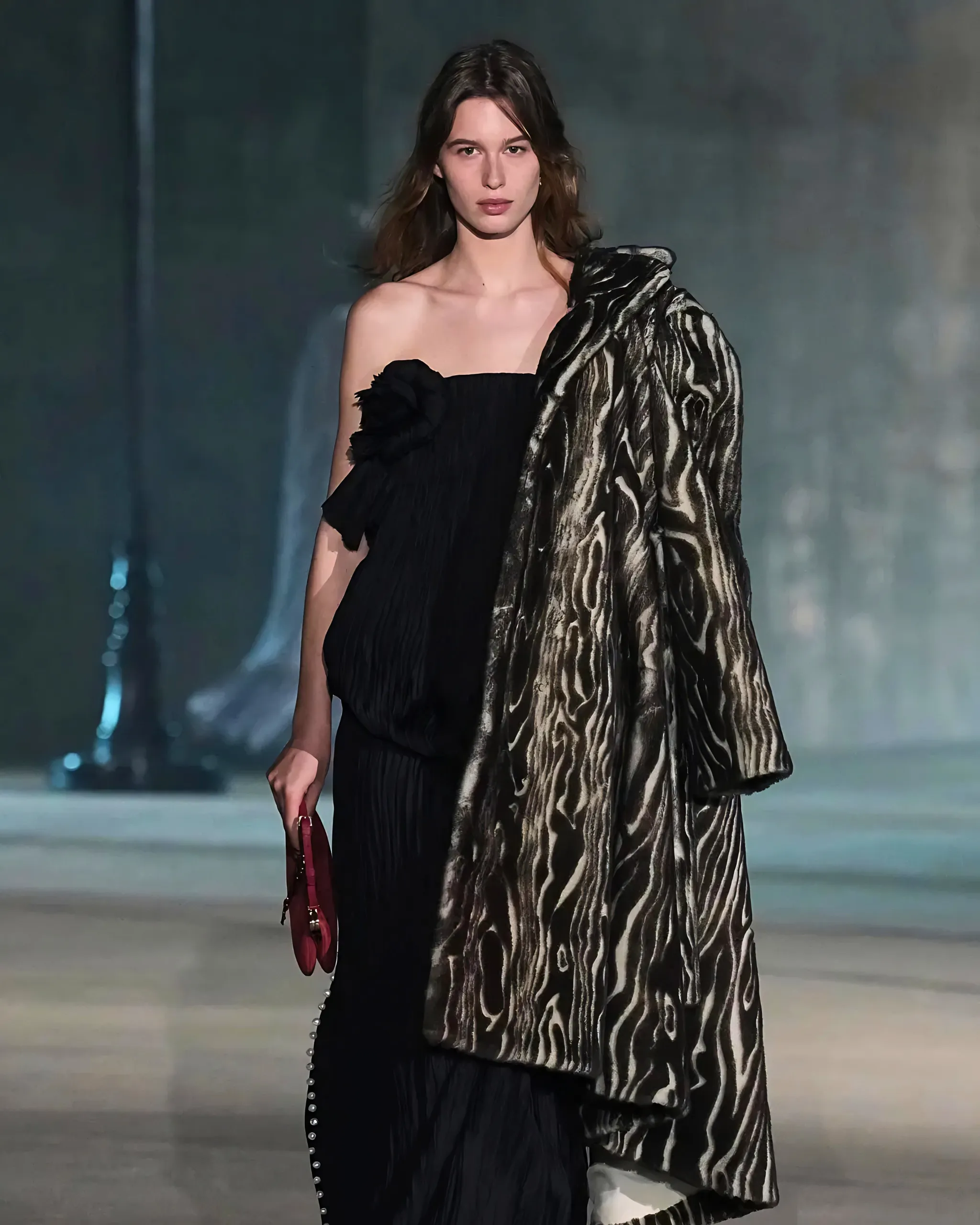 Model walking on a runway in a black strapless dress with a zebra-patterned shawl draped over one shoulder, holding a red handbag