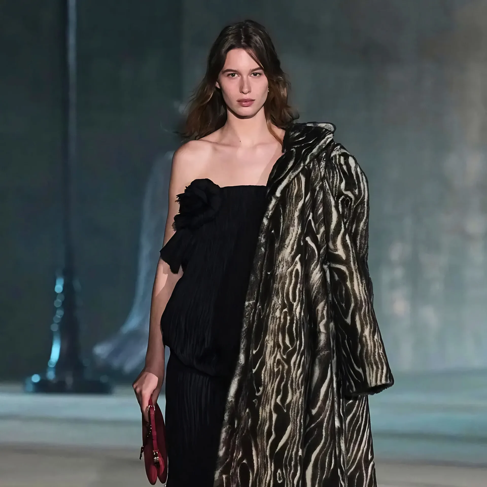Female model walking on a runway in a black off-the-shoulder dress with a zebra-pattern shawl, holding a red handbag.