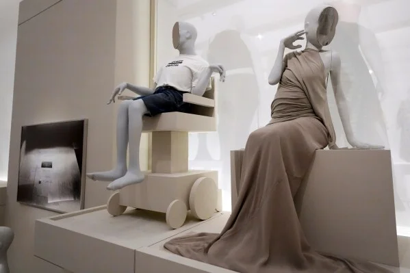 Mannequin display: a child-like figure sits on a cream stroller while a tall mannequin in a beige draped gown sits nearby on a pedestal.