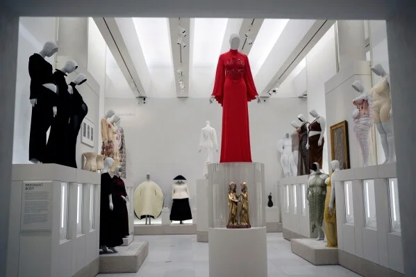 Museum display of mannequins in elegant dresses; a bright red gown stands on a pedestal at center.