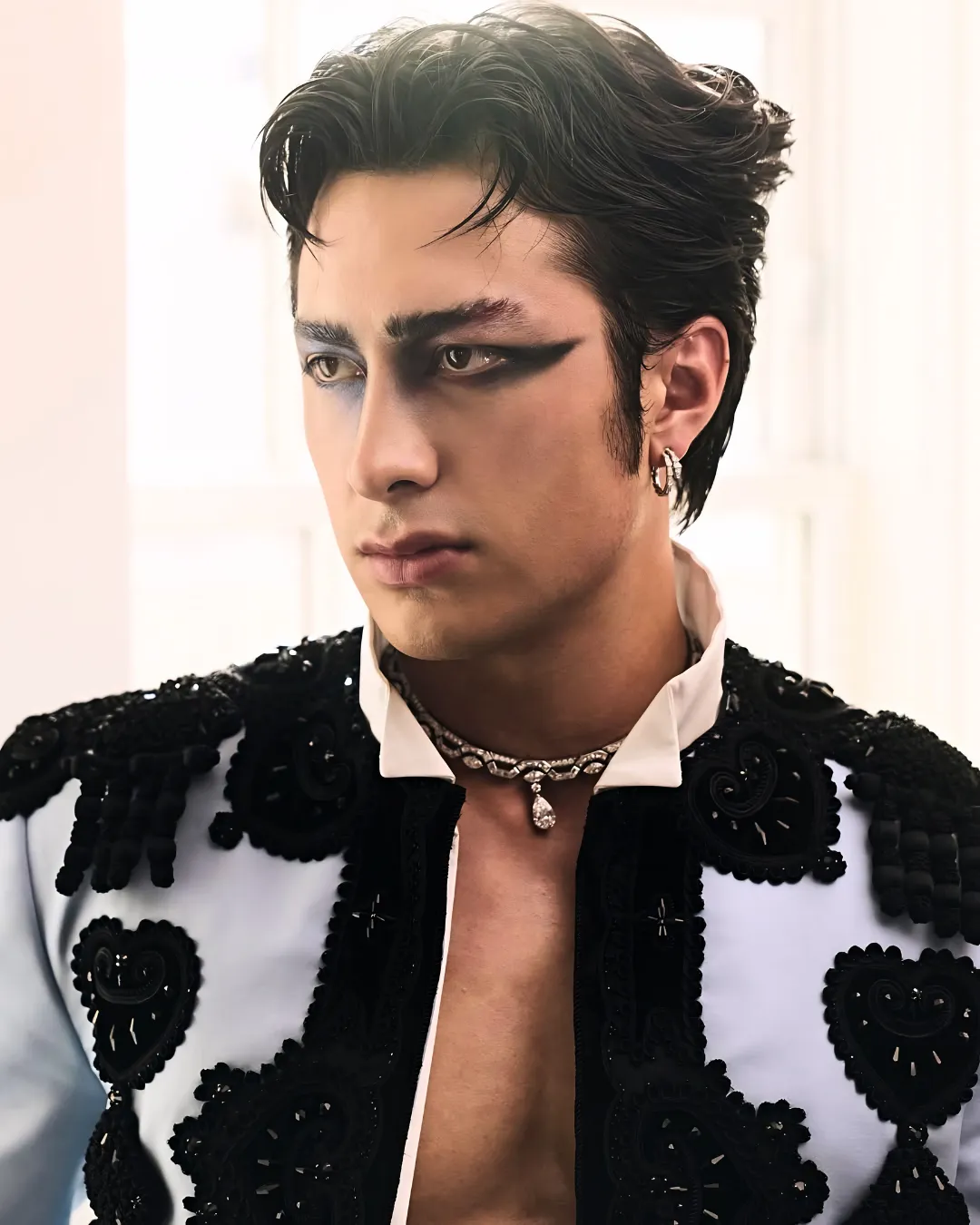Portrait of a young man with dramatic black eyeliner and styled hair, wearing a black-embellished white jacket, a jeweled necklace and earring, looking to the left indoors.