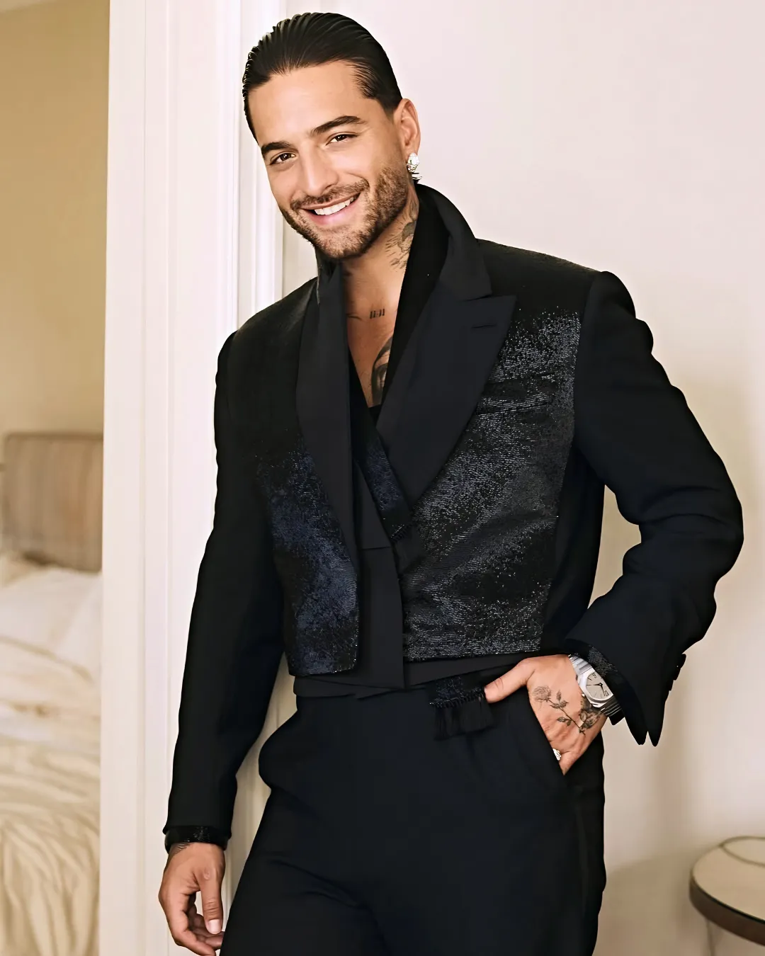 Smiling man in a black suit with a shimmering vest, leaning against a doorway.