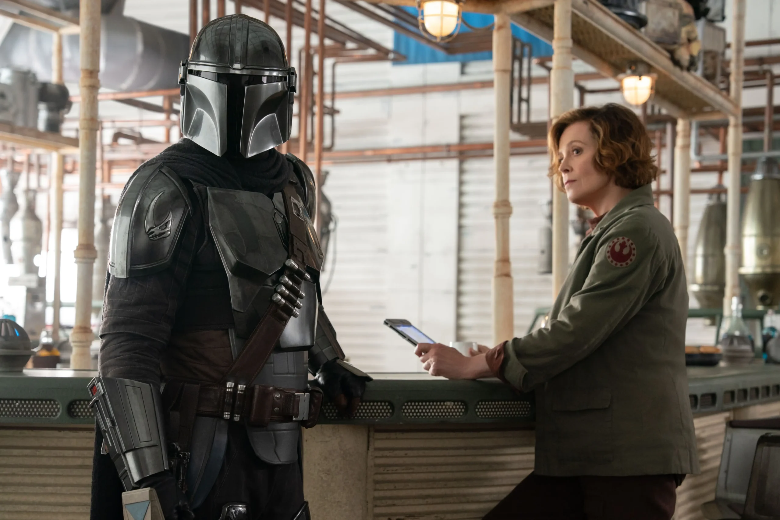 The Mandalorian stands in profile at a bar facing Colonel Ward, a New Republic officer played by Sigourney Weaver, who is holding a data pad.