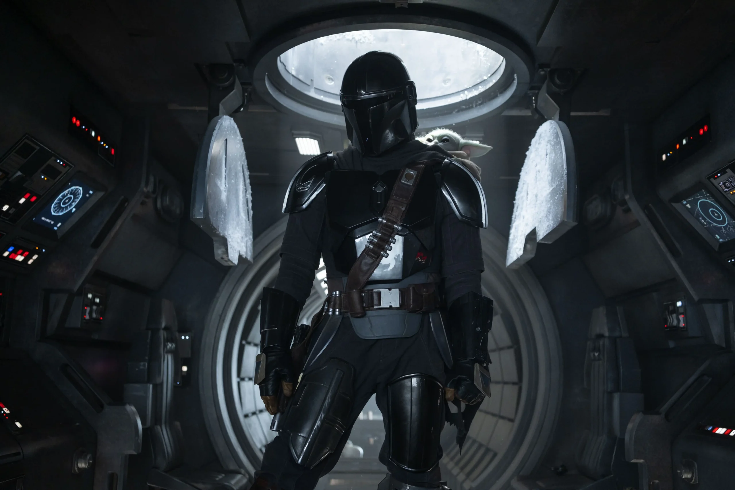The Mandalorian, clad in gleaming silver and black armor, stands centrally inside a dark, high-tech spaceship cockpit while Grogu peeks over his shoulder.