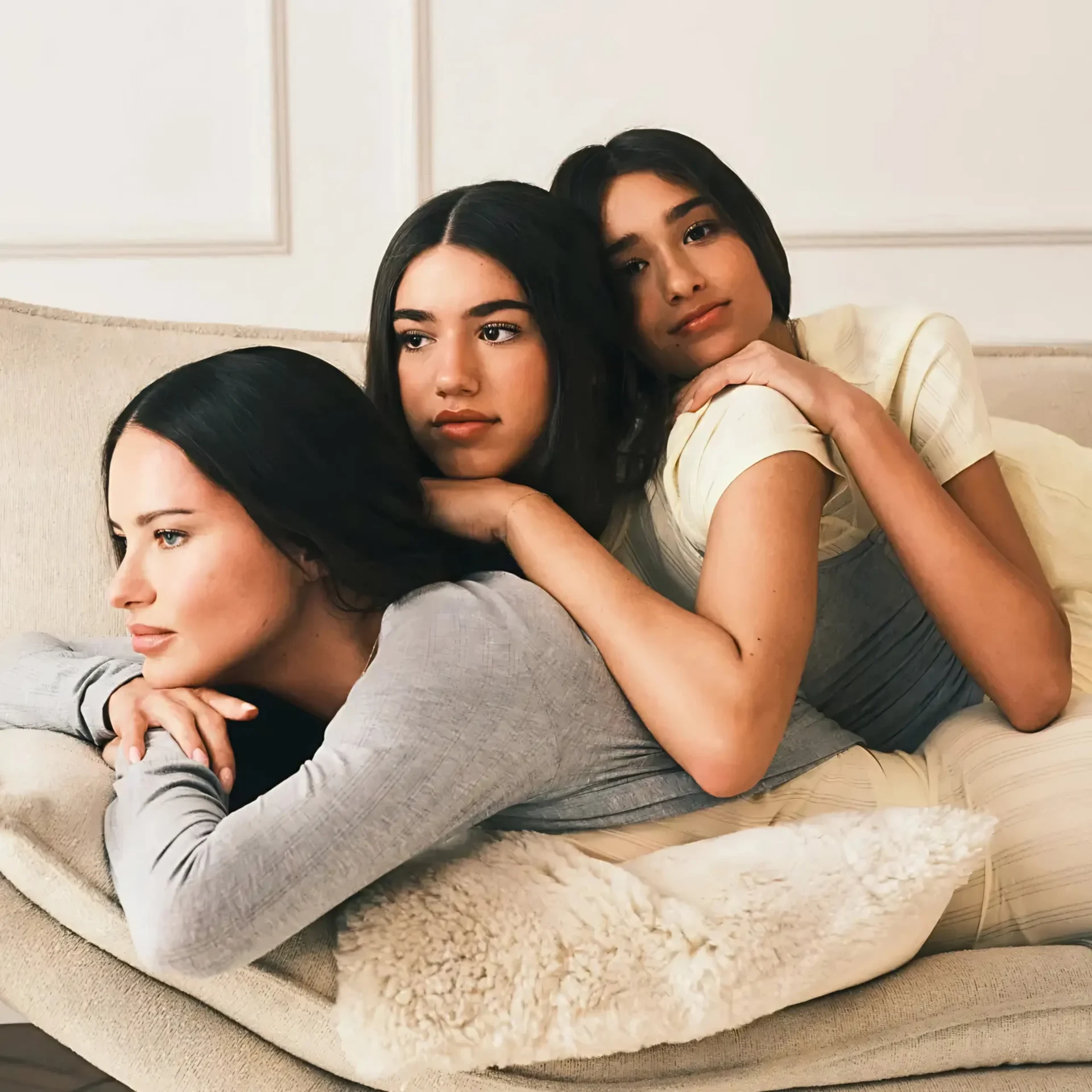 Three women cuddled together on a beige sofa, resting their heads on each other in a cozy living room setting.