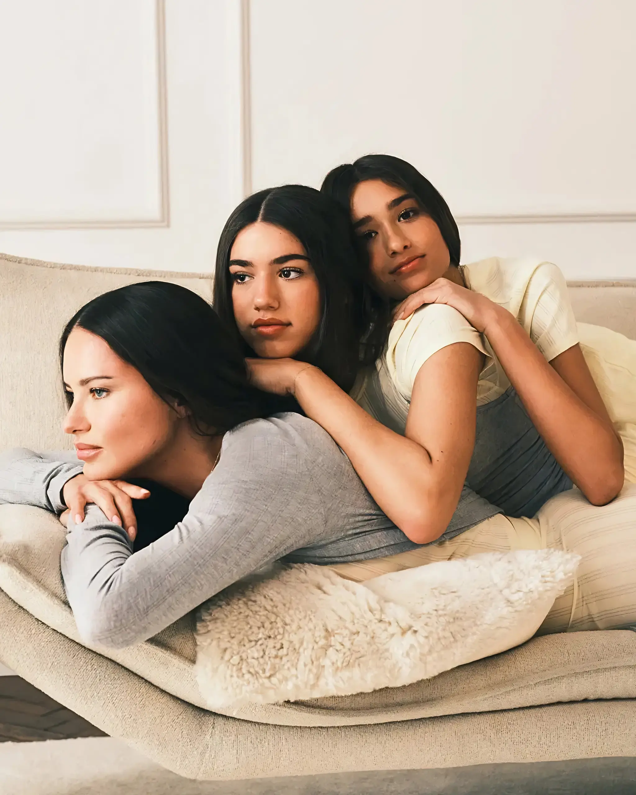 Three women cuddle together on a beige sofa, leaning against each other with calm expressions in a cozy living room.