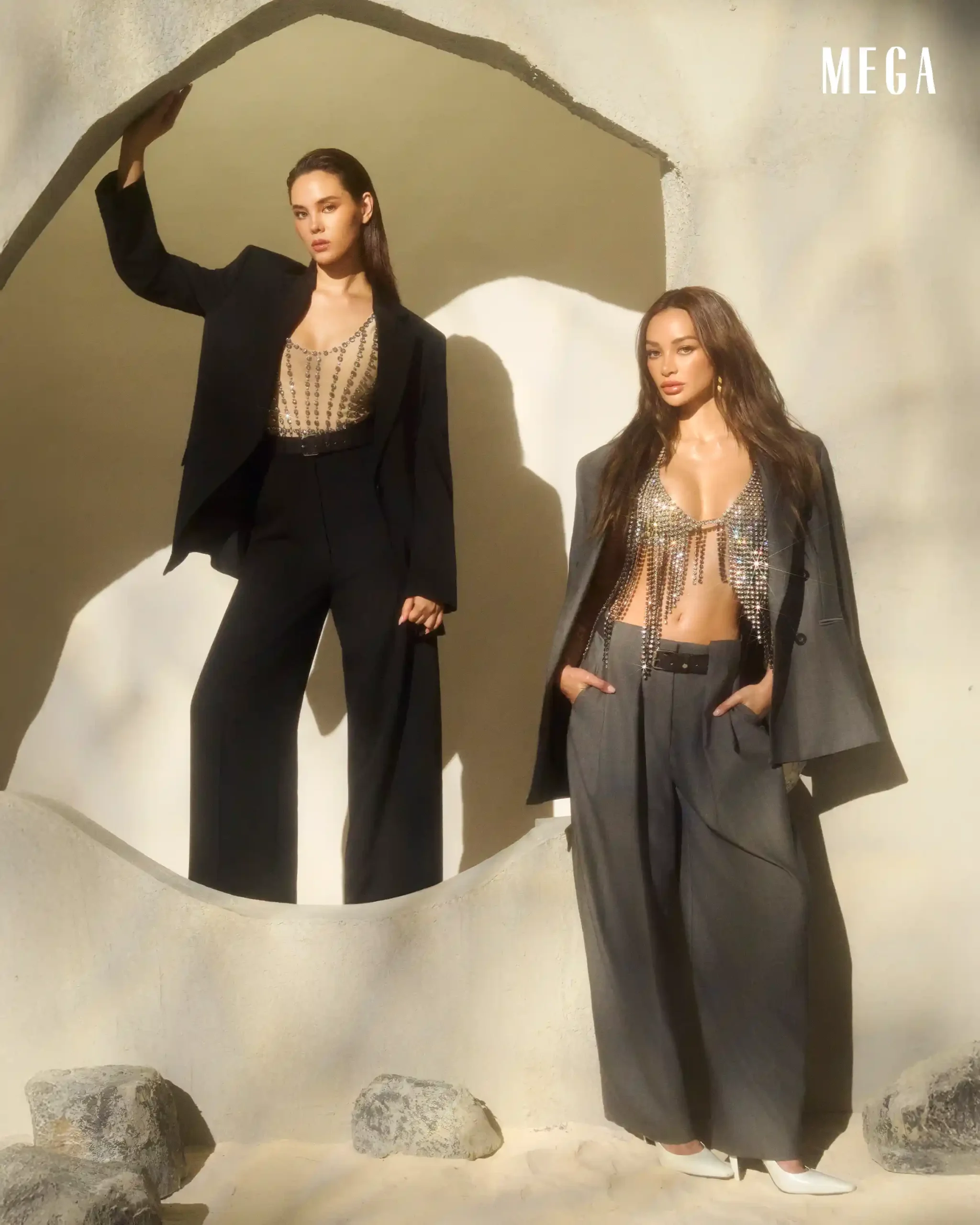 Two female models pose in a sunlit courtyard, wearing tailored black and gray suits with crystal embellishments; MEGA logo in the upper right.