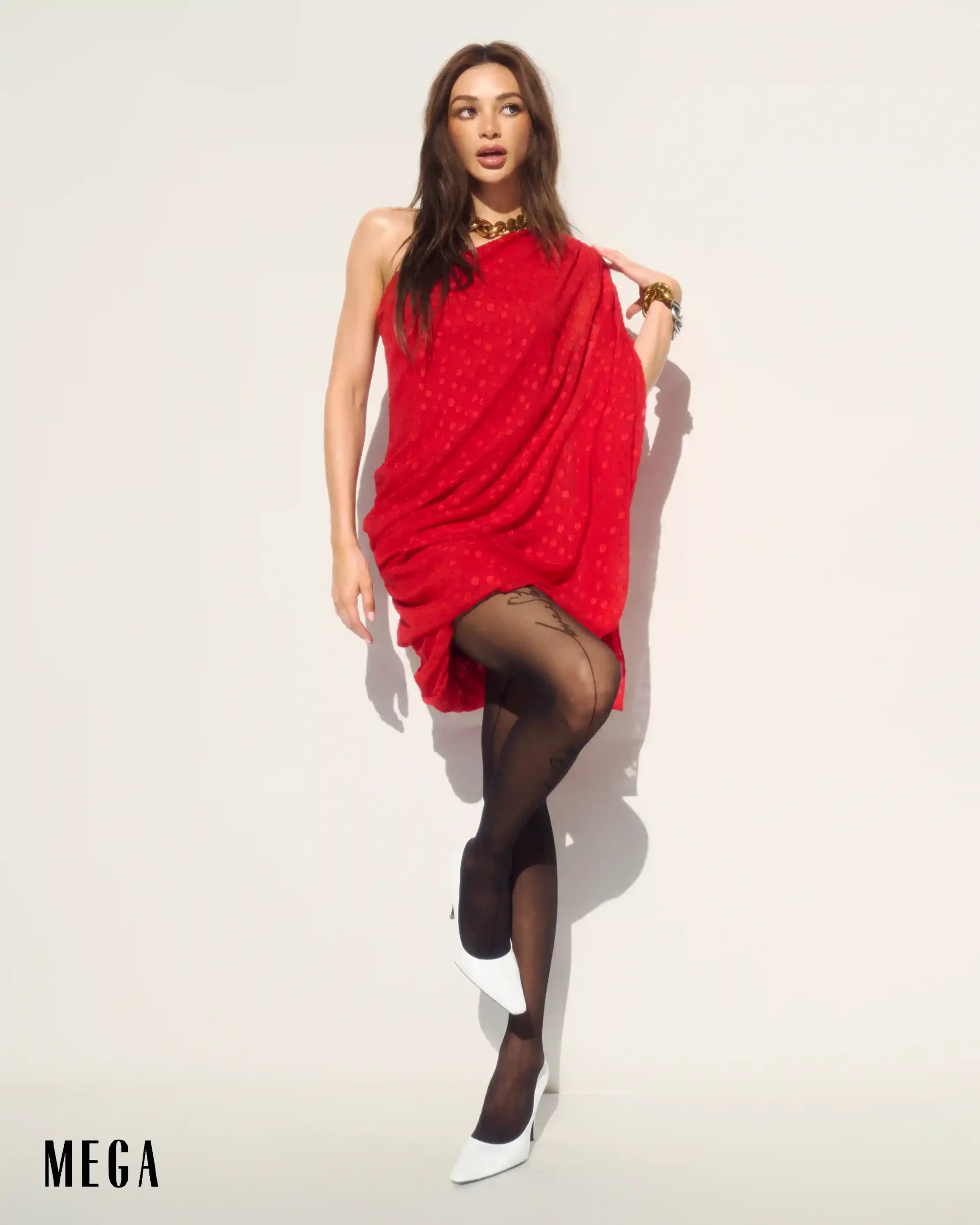 Model posing in a red draped dress with gold jewelry and white heels against a white studio backdrop.