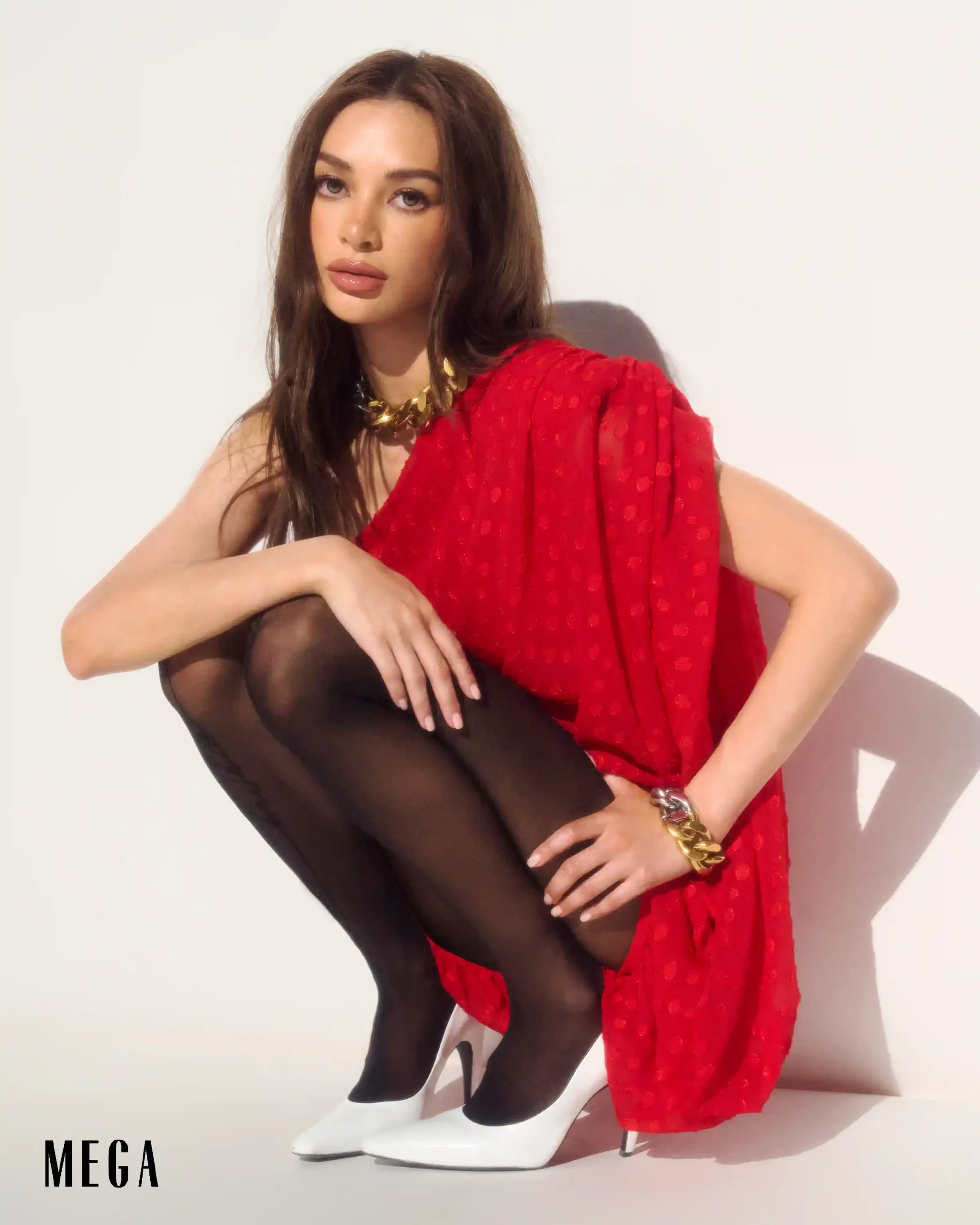 Model seated against a white background wearing a red sheer garment over black tights and white high heels, with a gold chunky necklace; MEGA logo bottom-left.