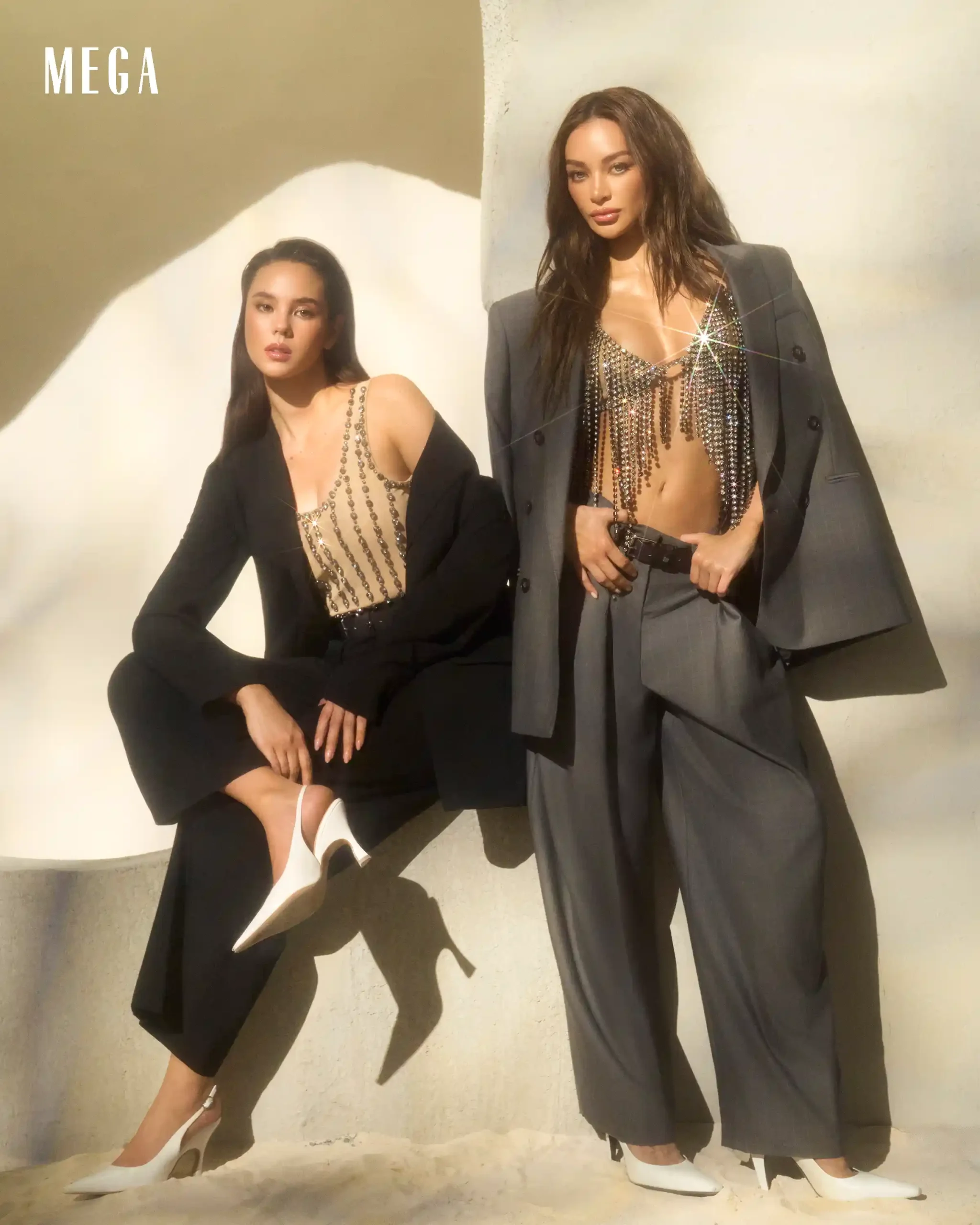 Two fashion models pose against a pale wall; one seated in a black suit with a crystal-beaded top, the other in an oversized gray suit with fringe details and white heels.