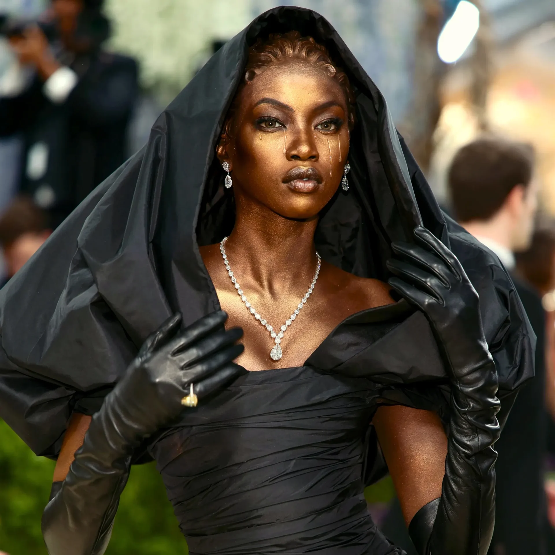 Close-up of a woman wearing a black satin gown and veil, with diamond earrings and a teardrop necklace, tears running down her cheeks against a blurred outdoor background.