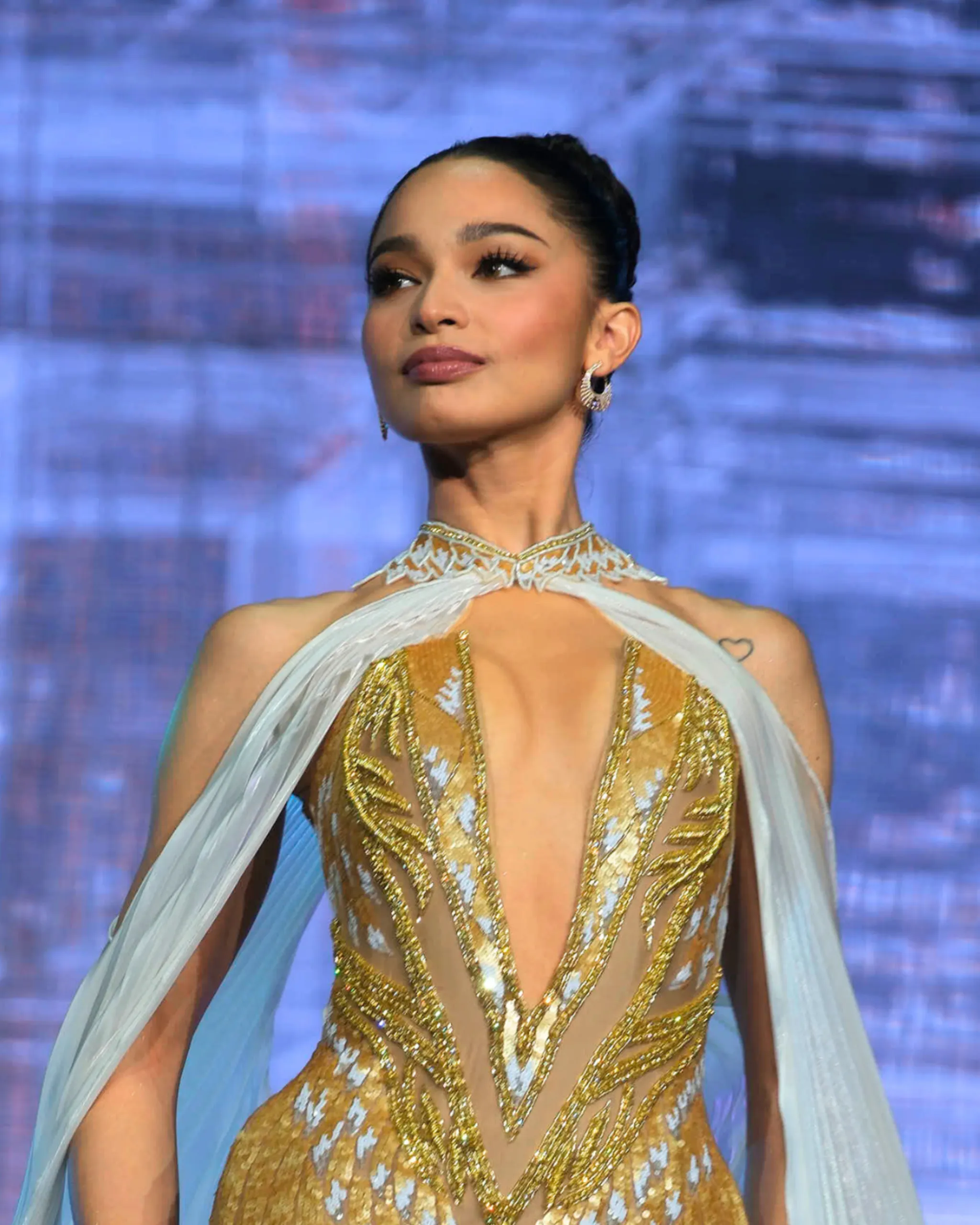 Portrait of a woman with dark hair in a gold sequined dress and sheer cape on a blue-lit stage, looking to the side.