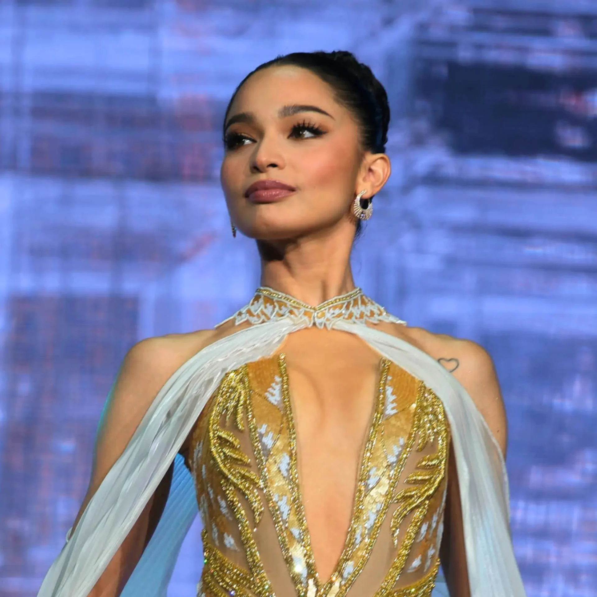 Woman wearing a gold sequined outfit and earrings, looking to the side on a blue backdrop stage.