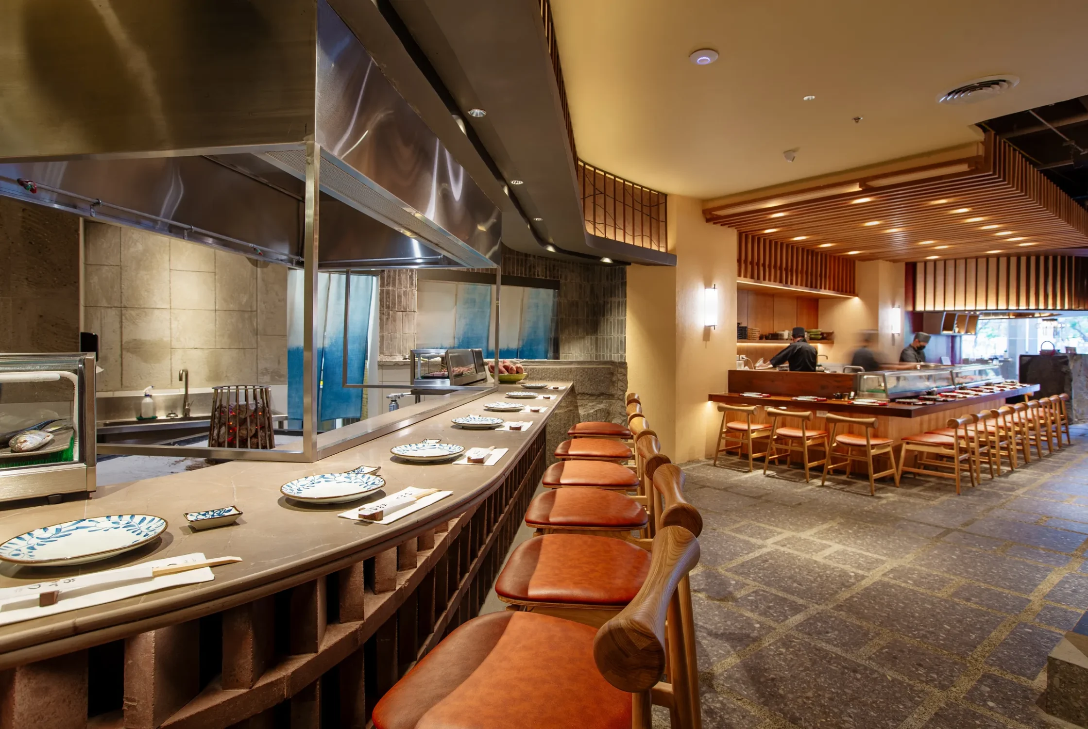 OGAWA BGC Sushi bar with a long counter and orange-brown stools, open kitchen behind stainless steel grills and prep area, warm lighting.