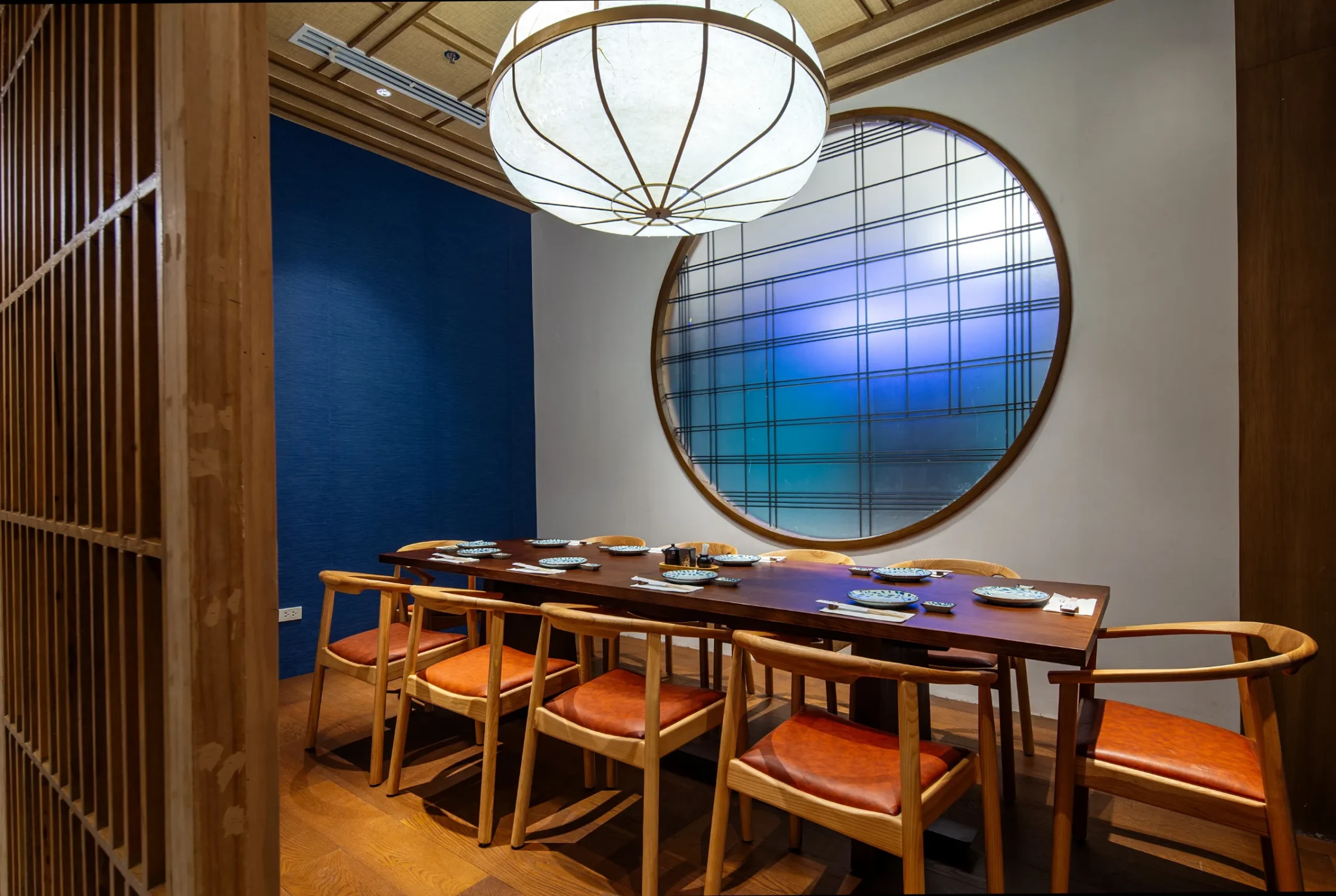 OGAWA BGC Wood-furnished dining room with a long table set for a meal, orange-cushioned chairs, and a large circular window with blue grid pattern on the wall behind.