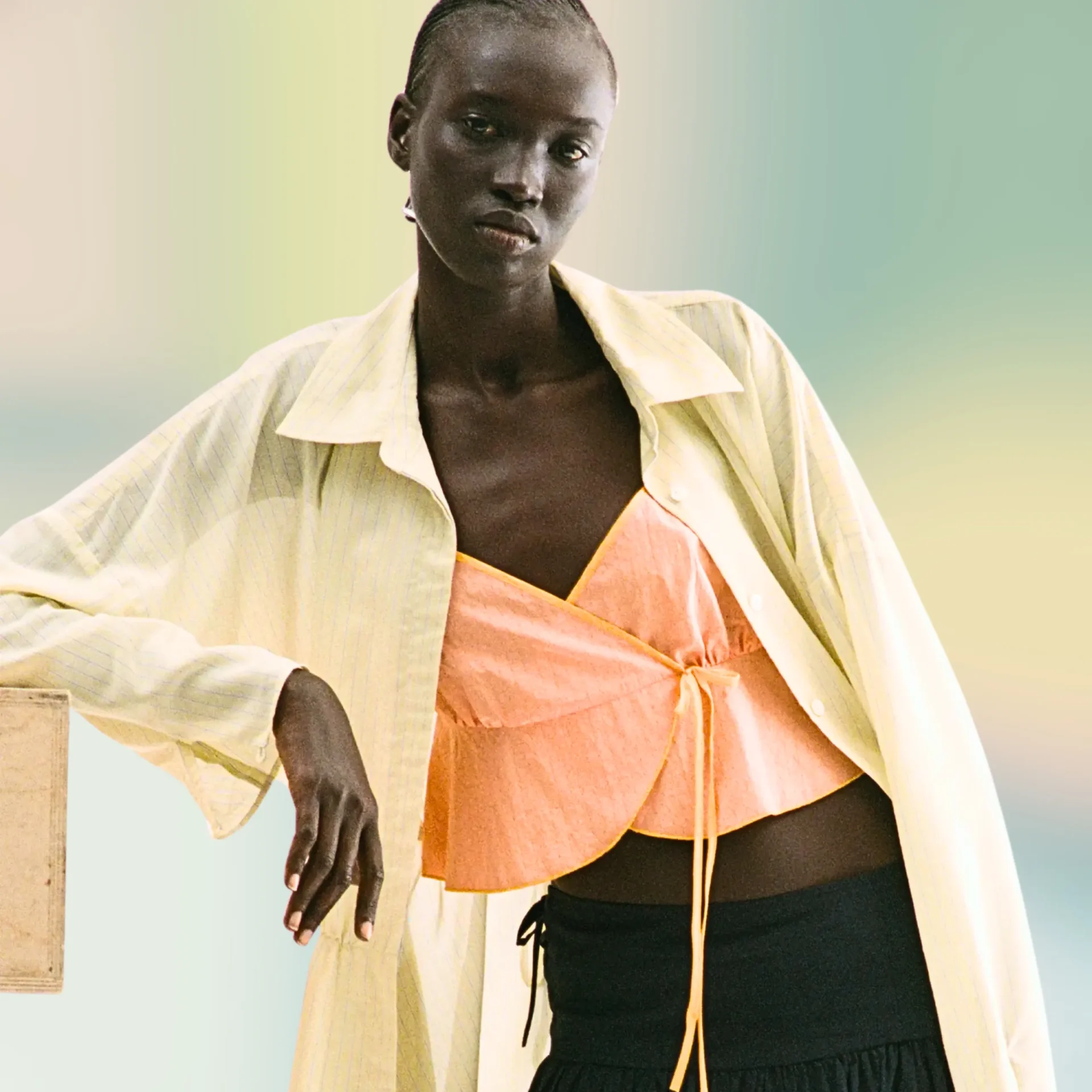 Person with dark skin wearing a pale striped shirt over a pink top, standing against a soft gradient background.