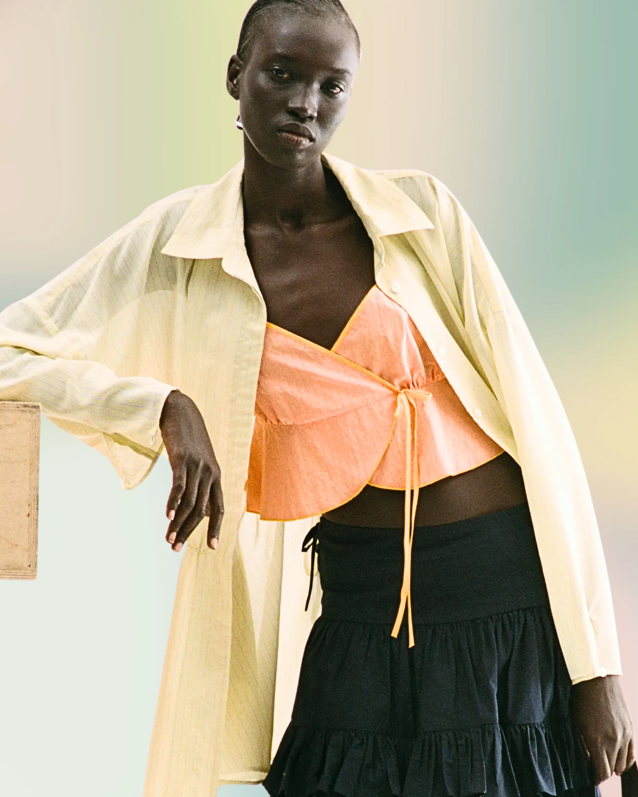 Person standing with one arm resting on a surface, wearing a peach top under an oversized striped shirt and a black ruffled skirt against a soft gradient background.