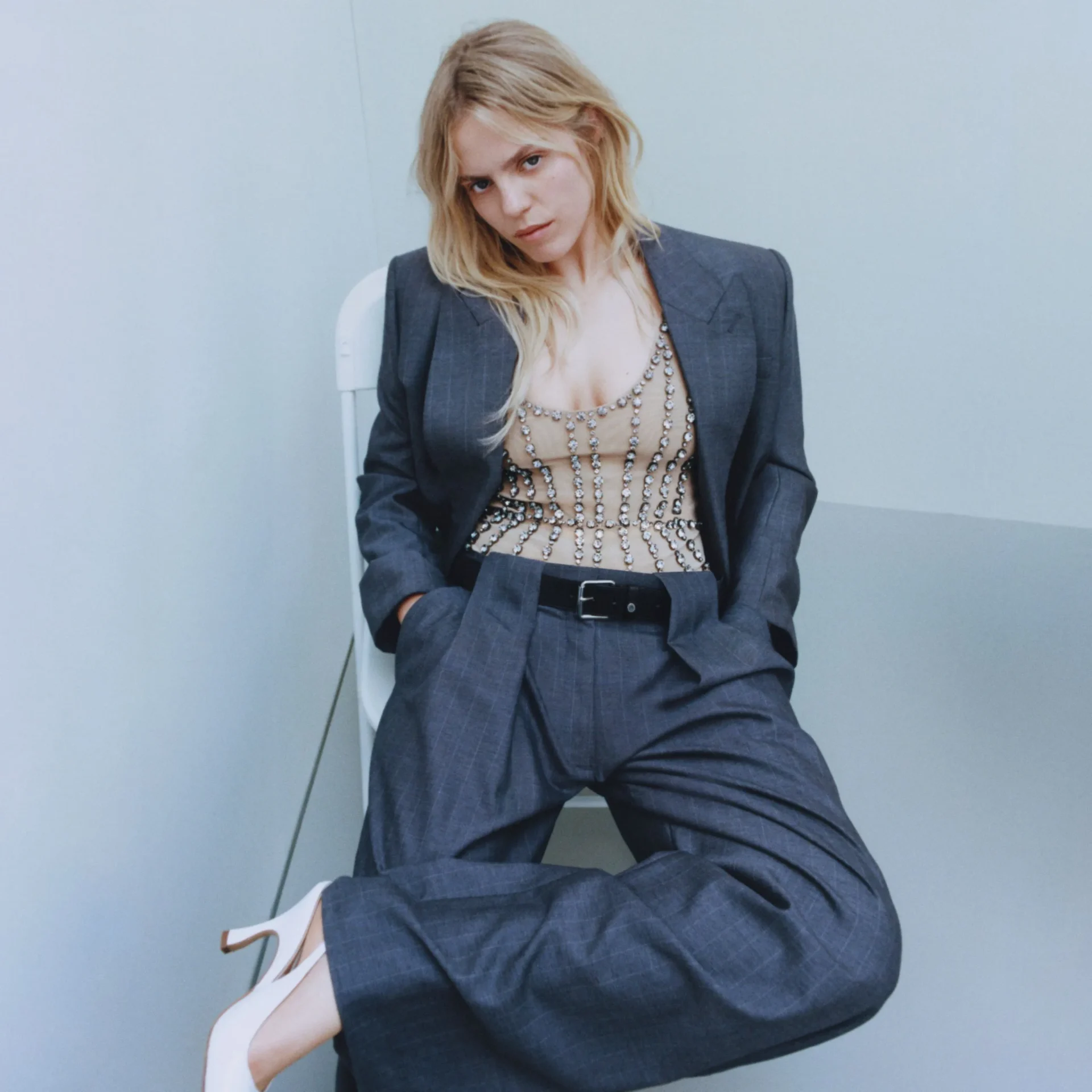 Woman in a dark pinstripe suit with a beige beaded top, seated against a pale blue wall, looking at the camera with a subtle expression.