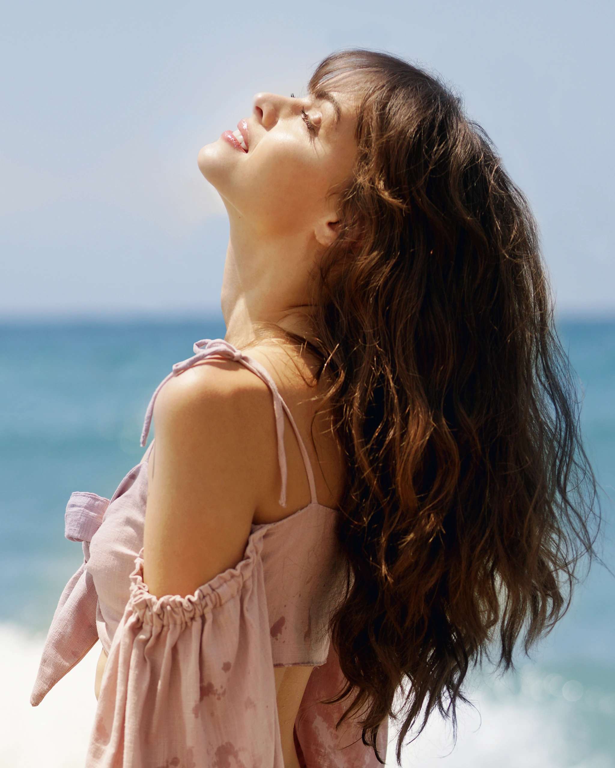 Profile of a woman with long brown hair in a pink dress, tilting her head back at the beach clear blue sky behind her