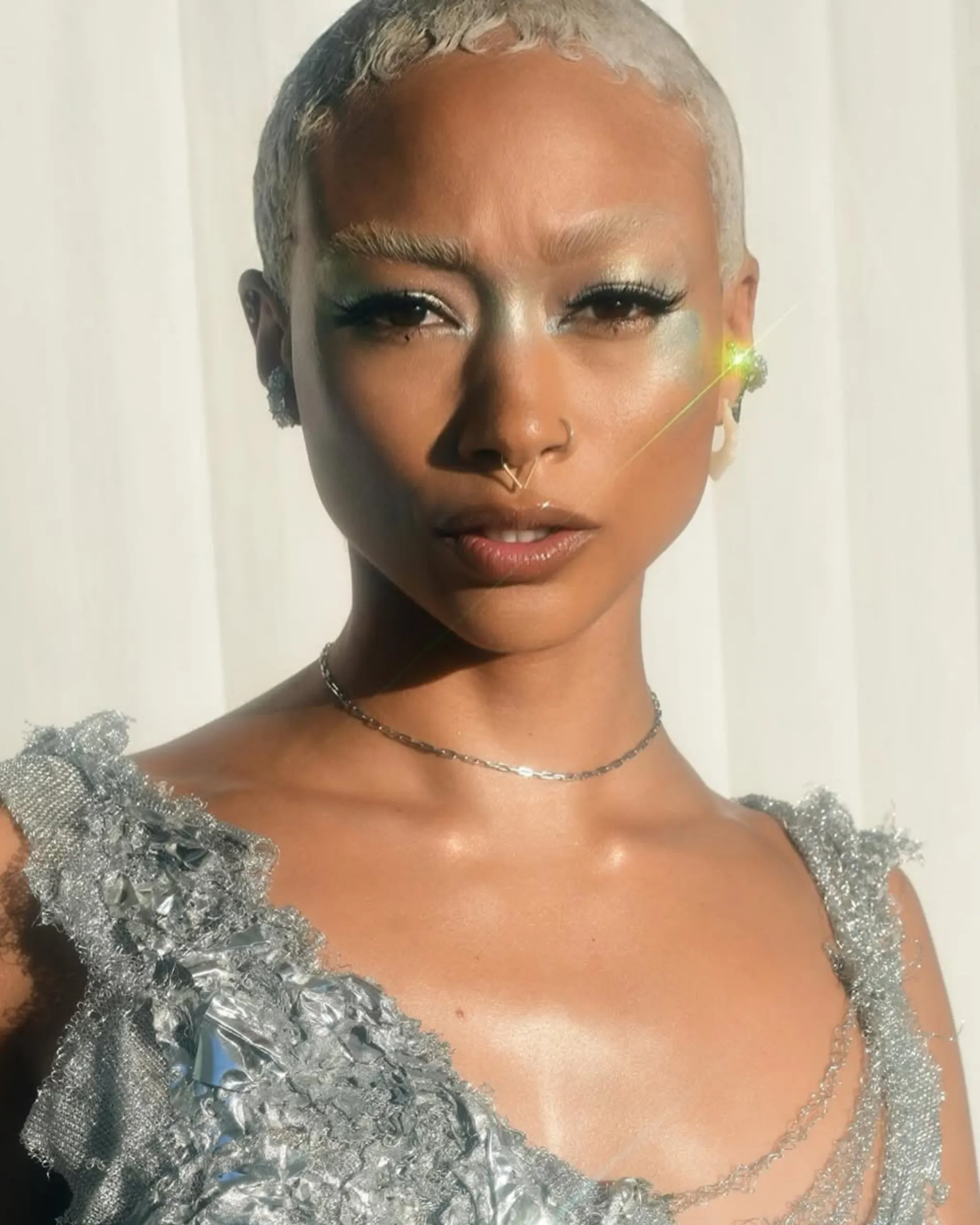 Close-up of a person with short platinum-blonde hair, shimmering silver makeup, and a textured metallic silver dress, facing the camera.