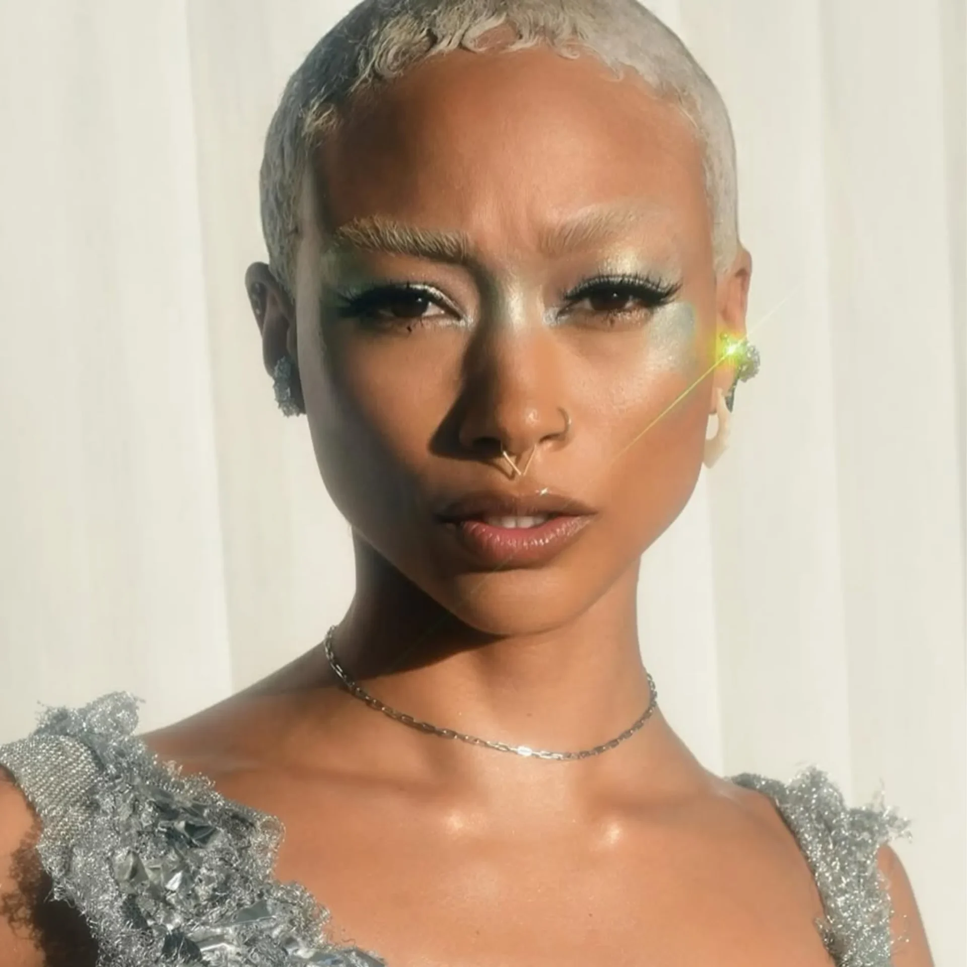 Close-up of a person with short platinum hair wearing a shimmering silver off-shoulder dress and a delicate necklace.
