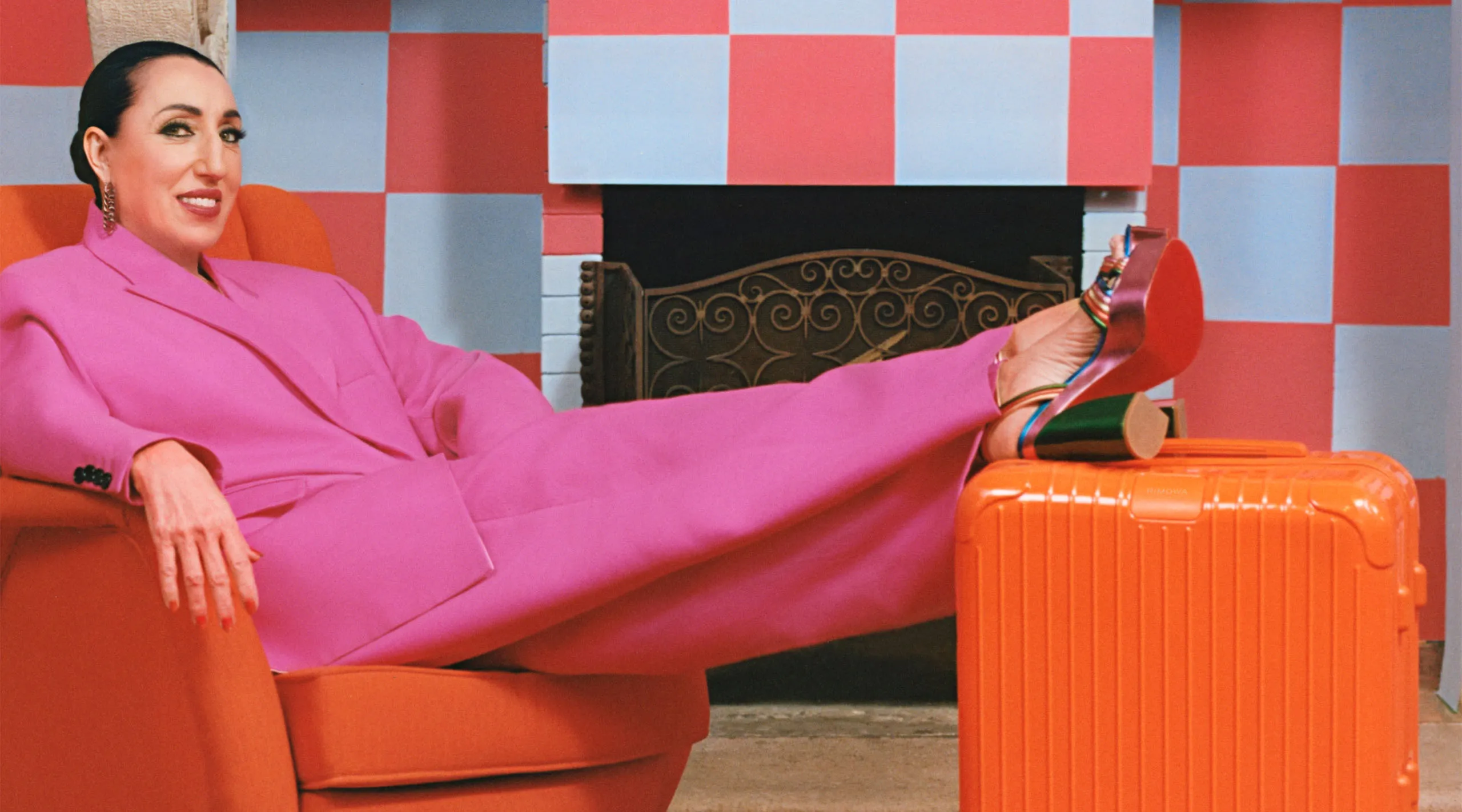 Actress Rossy de Palma, clad in bright magenta, lounging by her Orange Essential baggage