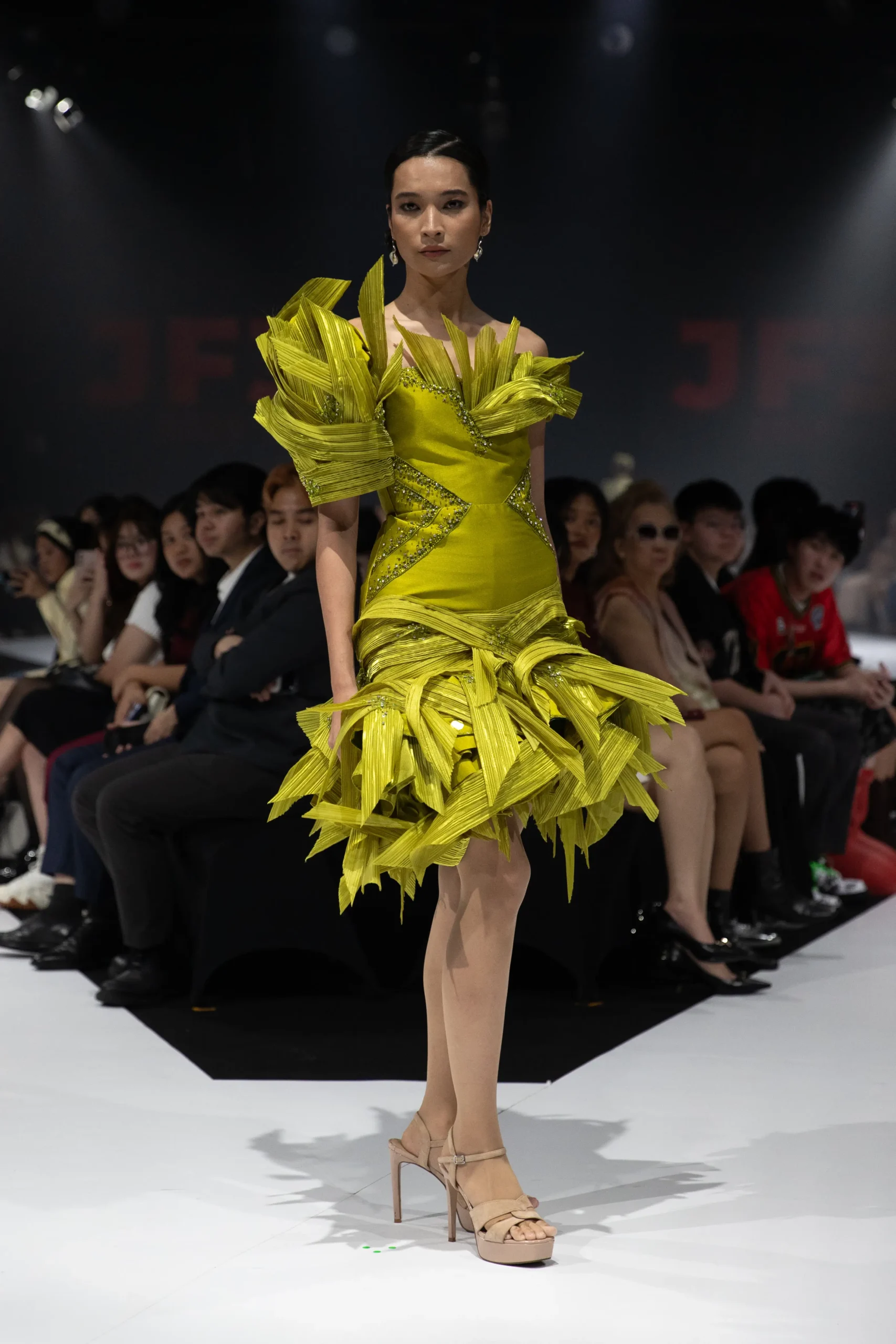 Model walks the runway in a vivid yellow gown with large feather-like shoulder details and geometric embellishments, audience in the background.