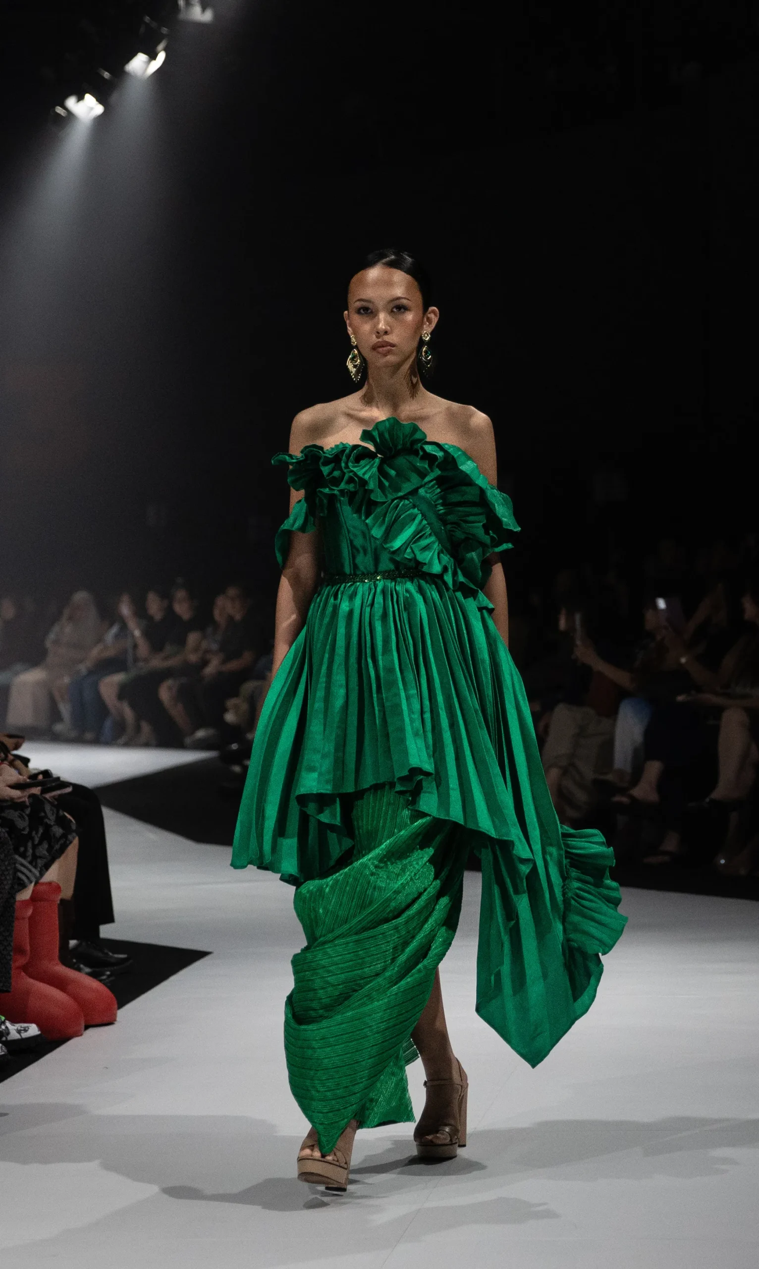 Model walks a fashion runway in a vivid emerald green off‑shoulder gown with large ruffles, under spotlight with a dark background.