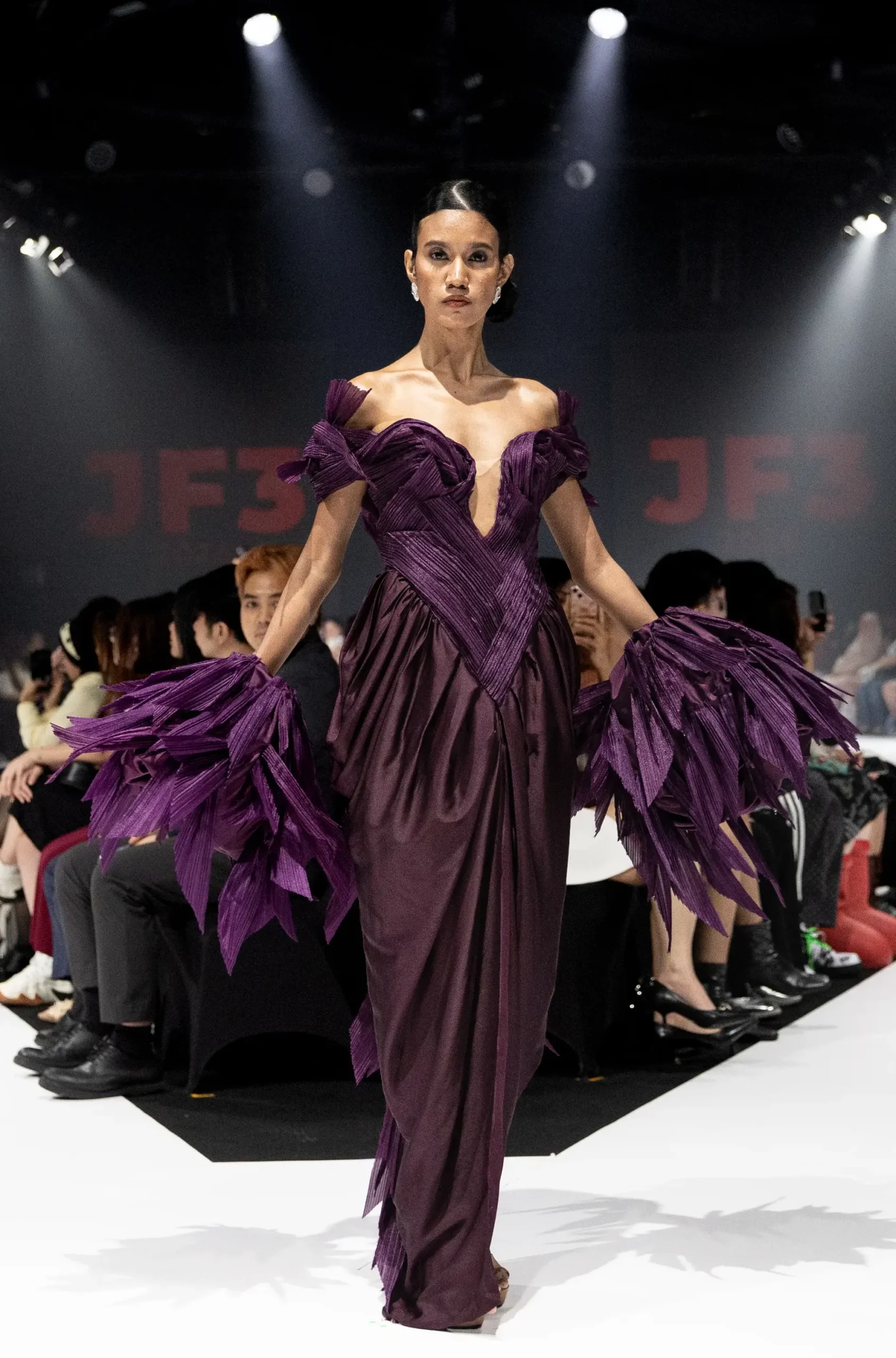 Model walks the runway in a deep purple gown with off-shoulder, pleated bodice and feathered sleeves under spotlight.
