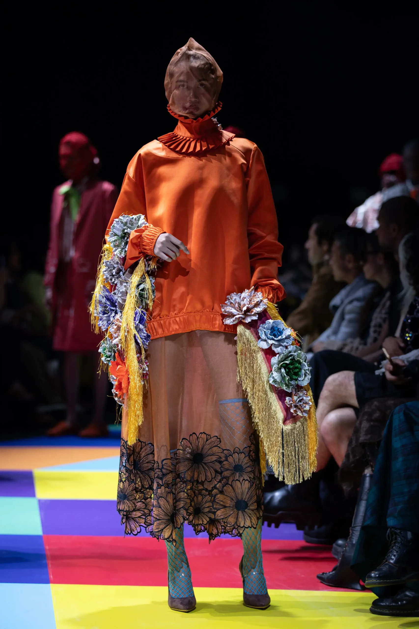 Model on a colorful runway wearing an orange dress with a ruffled collar, a hooded headpiece, and floral-printed hem; turquoise fishnet stockings and brown shoes visible.