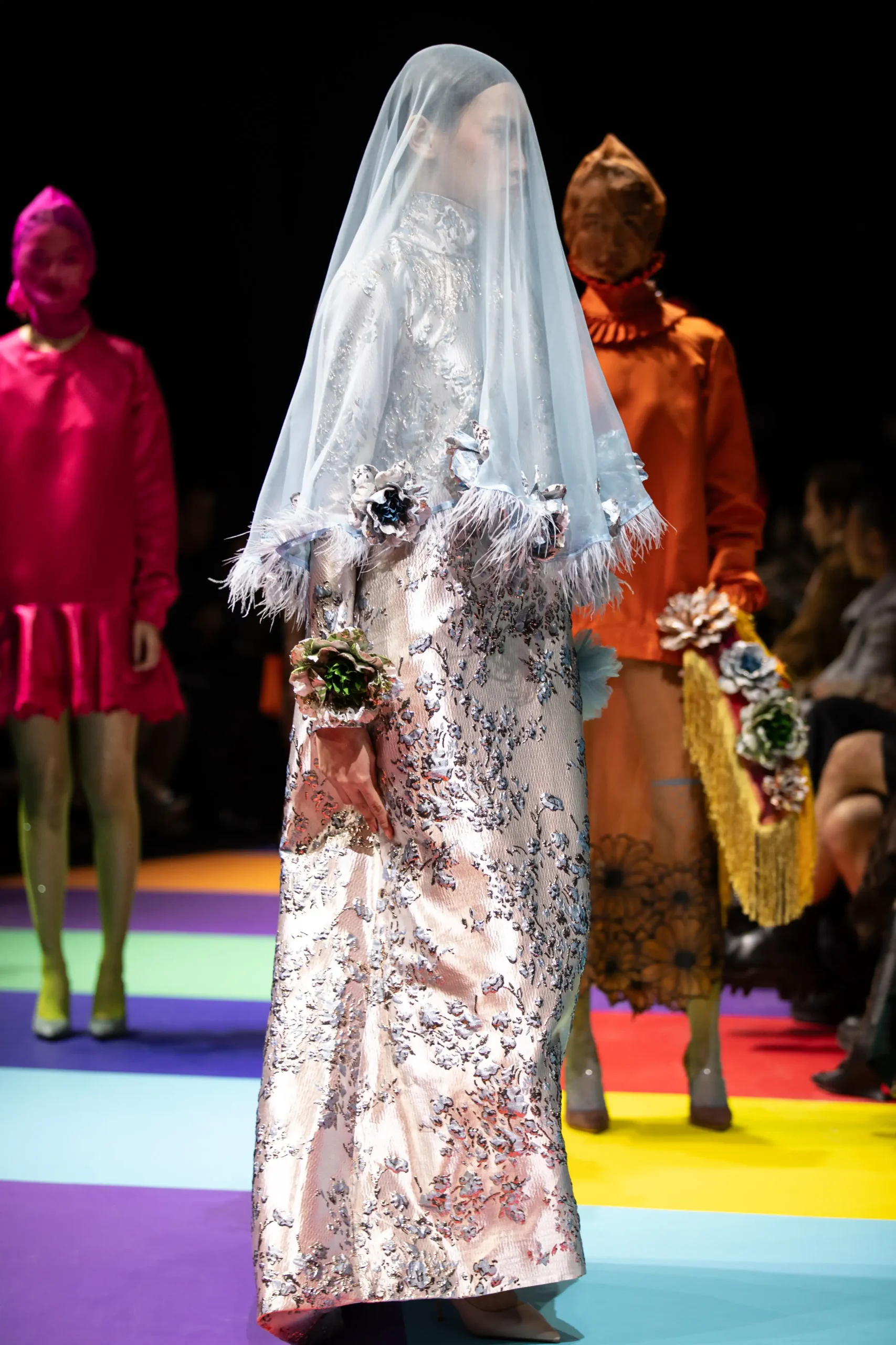 Model wearing a pale blue veil over an embroidered silver floral gown on a fashion runway, with bright pink and orange outfits in the background.