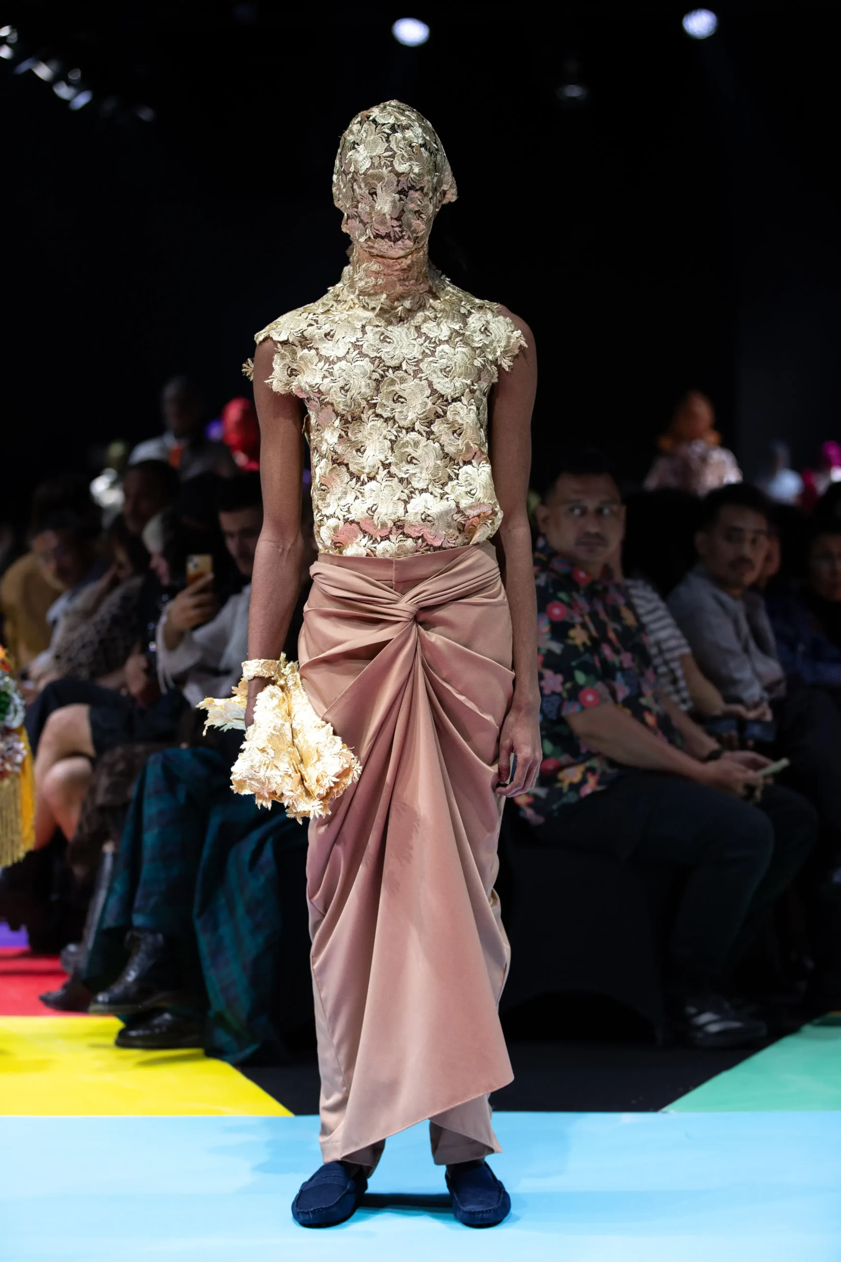 Model walks the runway in an ivory lace floral top that covers the head and shoulders, paired with a pink satin draped skirt, holding a cream bouquet.